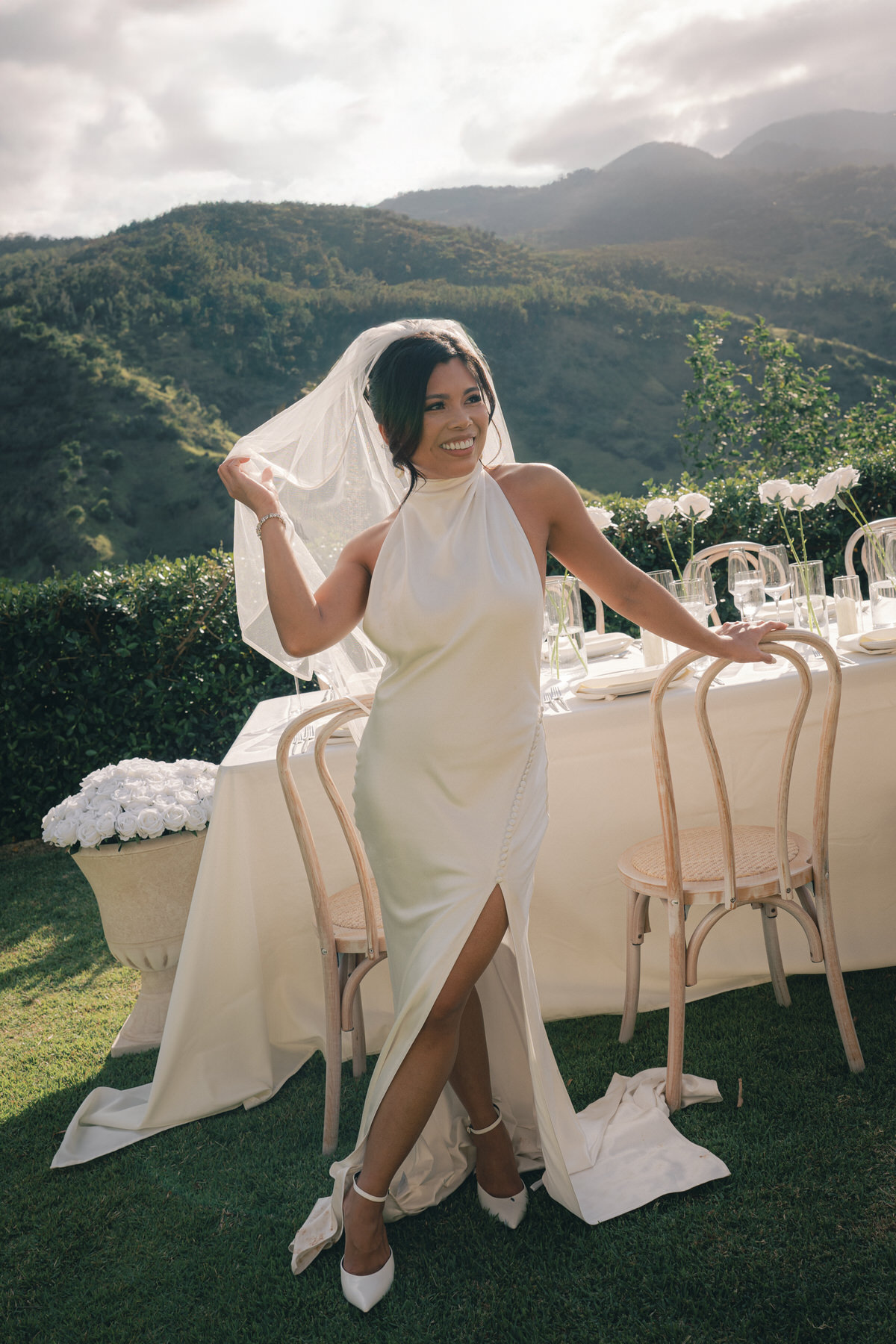 Bride in a white dress and veil poses outdoors with a scenic mountain backdrop. She stands next to a table set for an event, smiling and holding her veil.