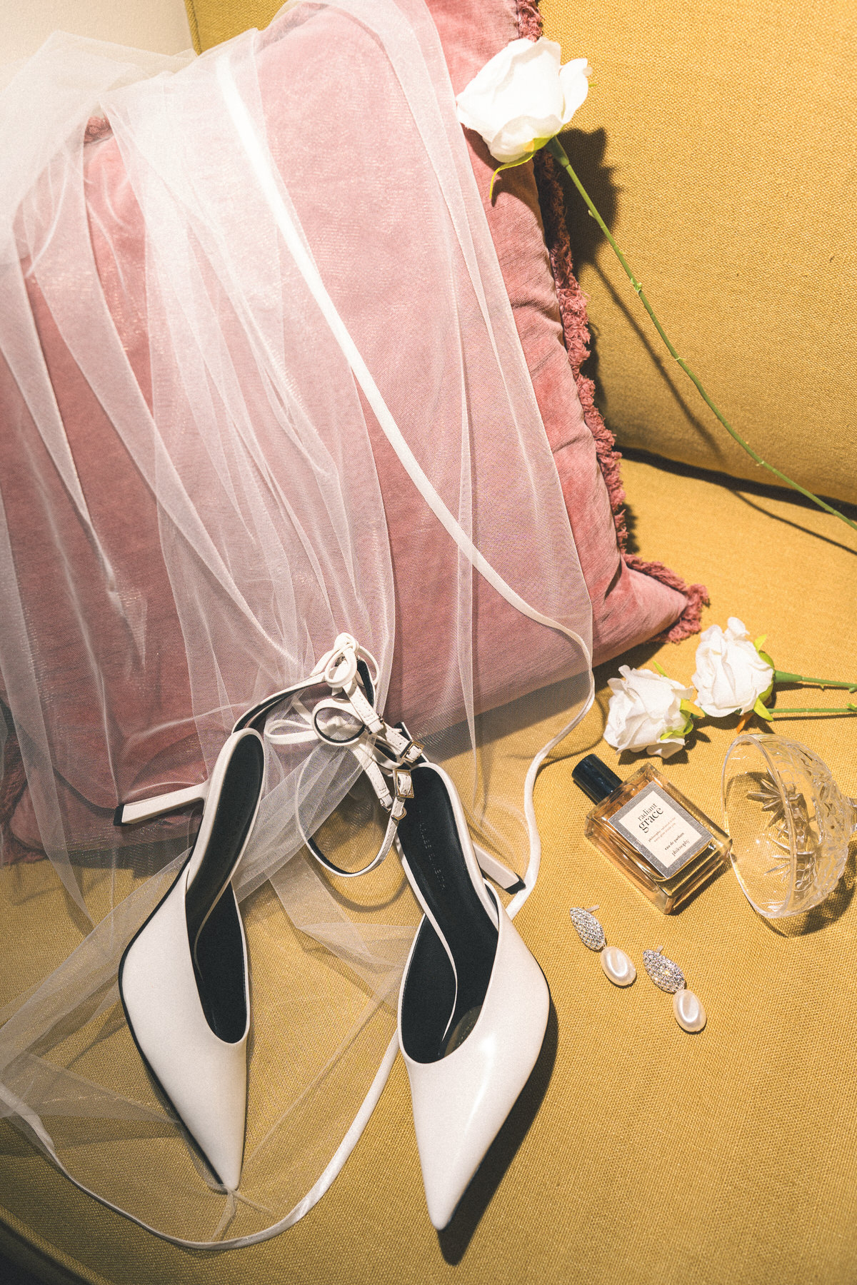 Bridal shoes, veil, perfume bottle, and earrings laid out on a yellow textured fabric with white roses.