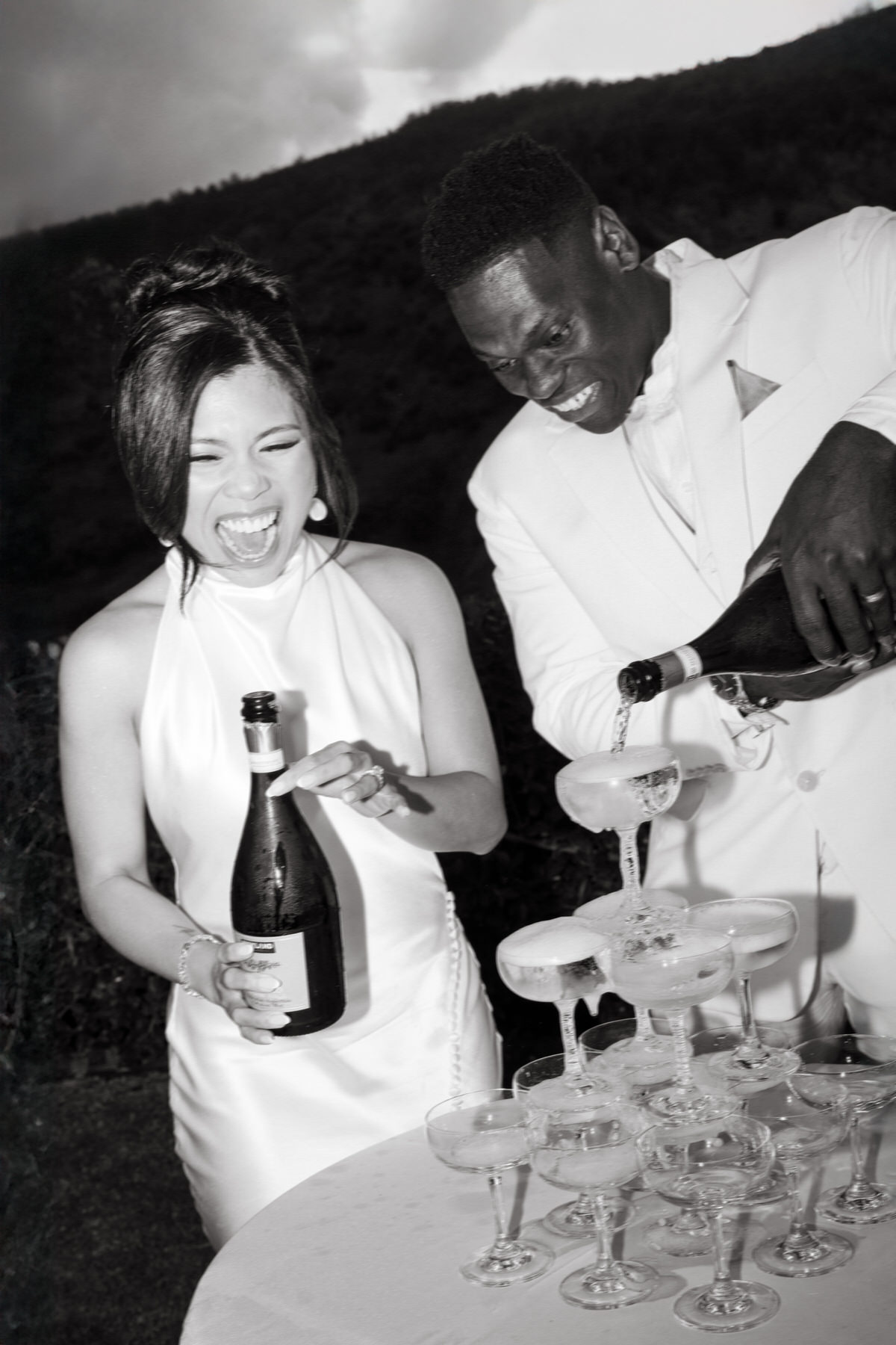 A couple in formal attire pours champagne into a tower of glasses outdoors. Both are smiling and appear joyful. The background shows a hillside. Image is in black and white.