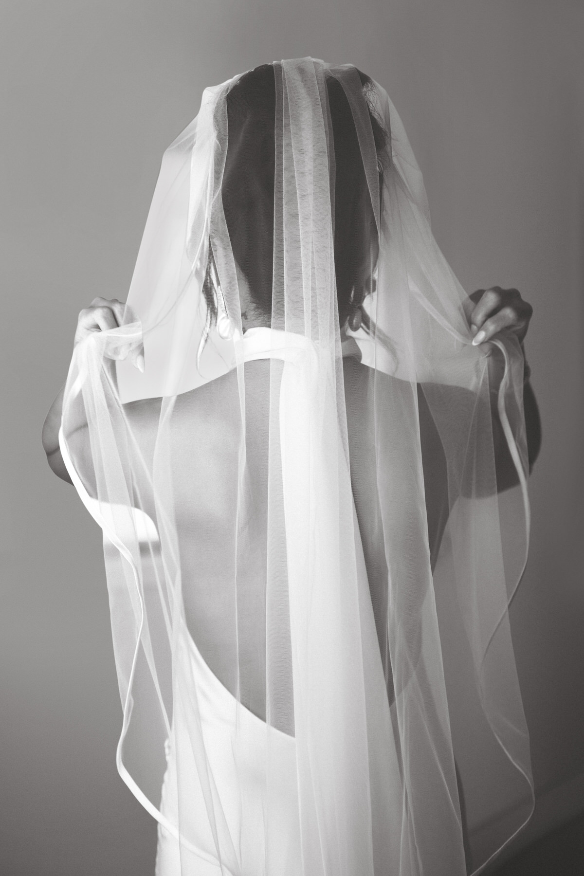 Black and white photo of a bride from behind, wearing a sheer veil over a backless dress, creating a serene and timeless moment.