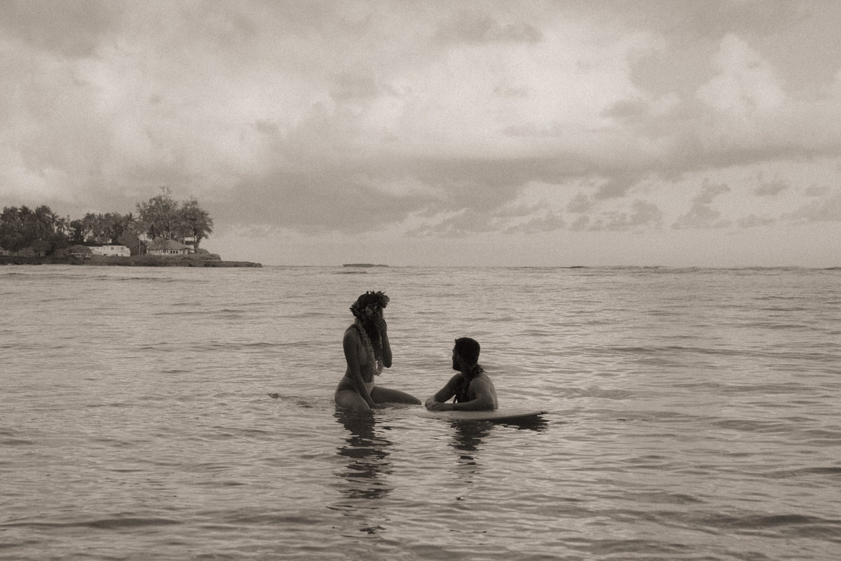 A black-and-white image of a couple in the ocean, enjoying a quiet moment. The woman, in a swimsuit, stands knee-deep in the water, while the man is seated on a floating surfboard, gazing at her, with a calm and expansive sky in the background at Kalākaua Avenue