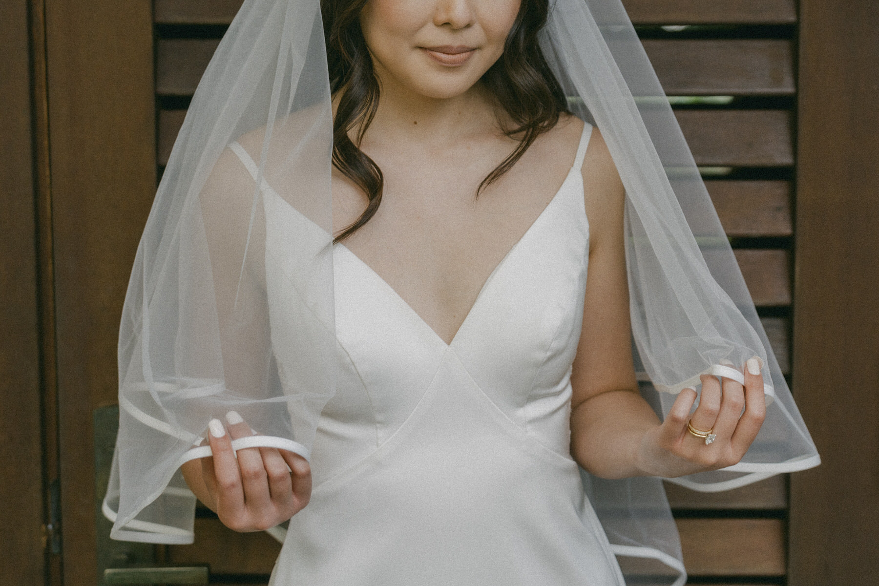 A bride in a simple white gown holds her veil up, showing her rings and delicate nails. The image captures her grace, offering a timeless moment in a Photography Guide in Kapolei.