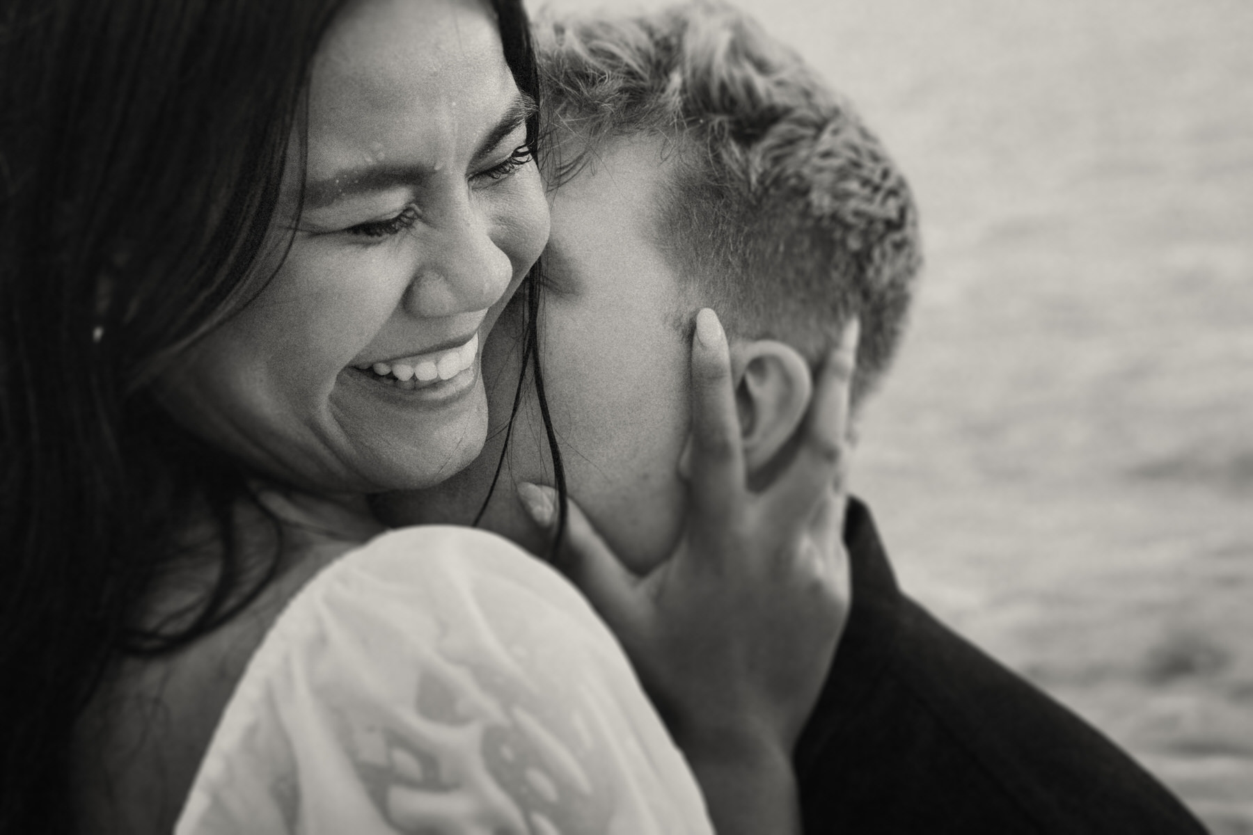 A close-up of a couple kissing at the beach, both laughing joyfully, capturing the love and energy shared between them. The ocean and sun create a beautiful backdrop for intimate beach photography in Kaneohe.