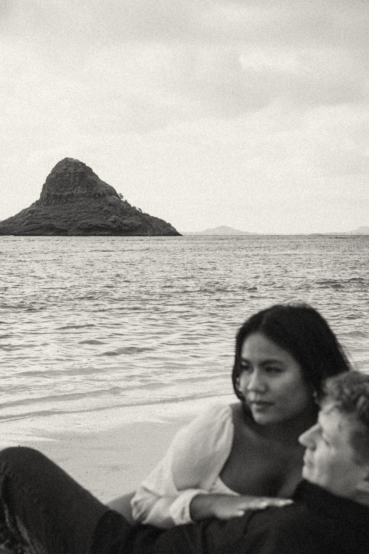 A couple relaxes by the ocean, lying together on the sand with Chinamans Hat in the background, a perfect spot for capturing intimate couple moments during a beach photography session in Kaneohe.