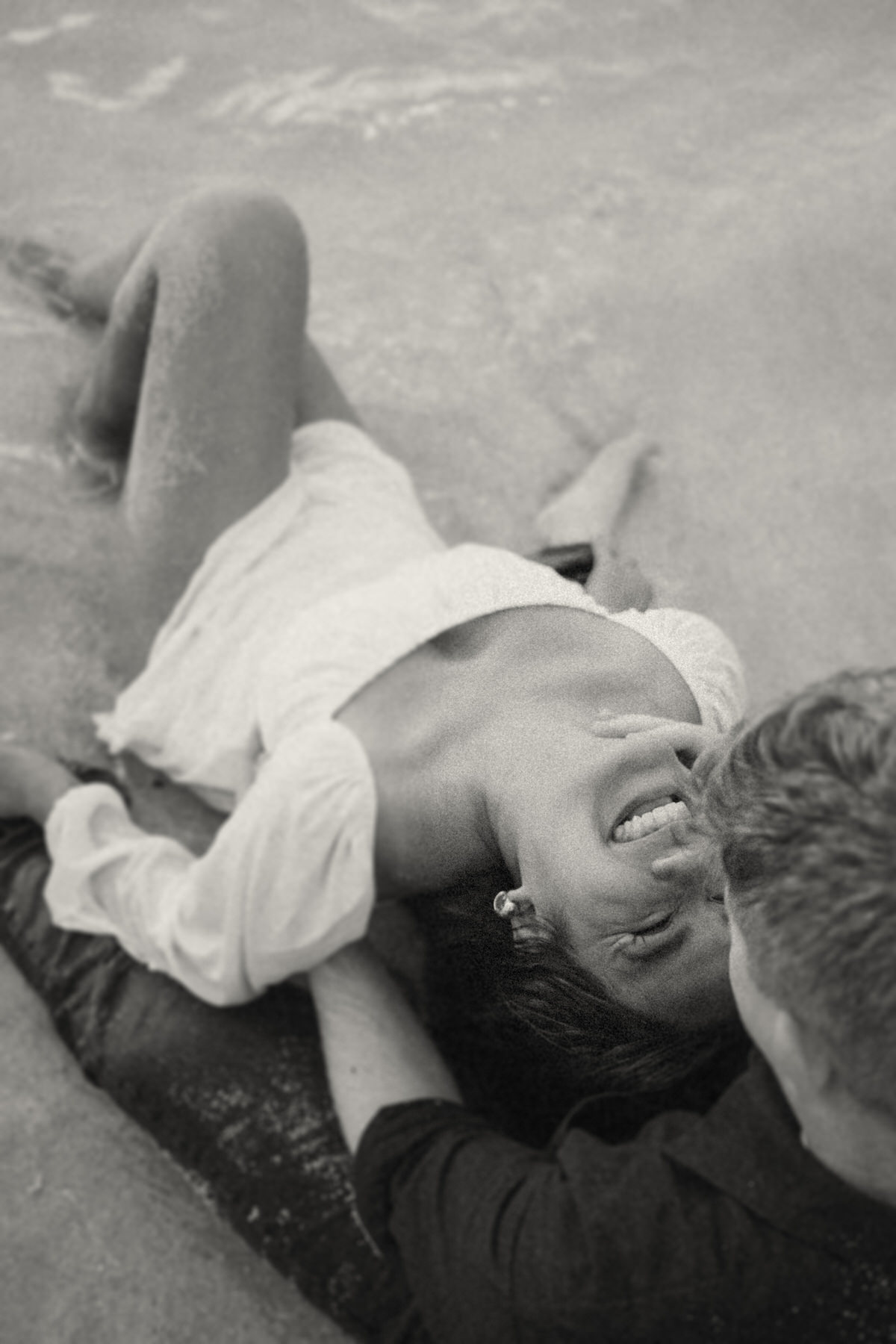 A couple shares a passionate kiss by the shore, with the woman laughing as she lays in her partner's arms. The moment is framed by the ocean and sky, reflecting the laid-back yet romantic vibe of a beach engagement shoot in Kaneohe.