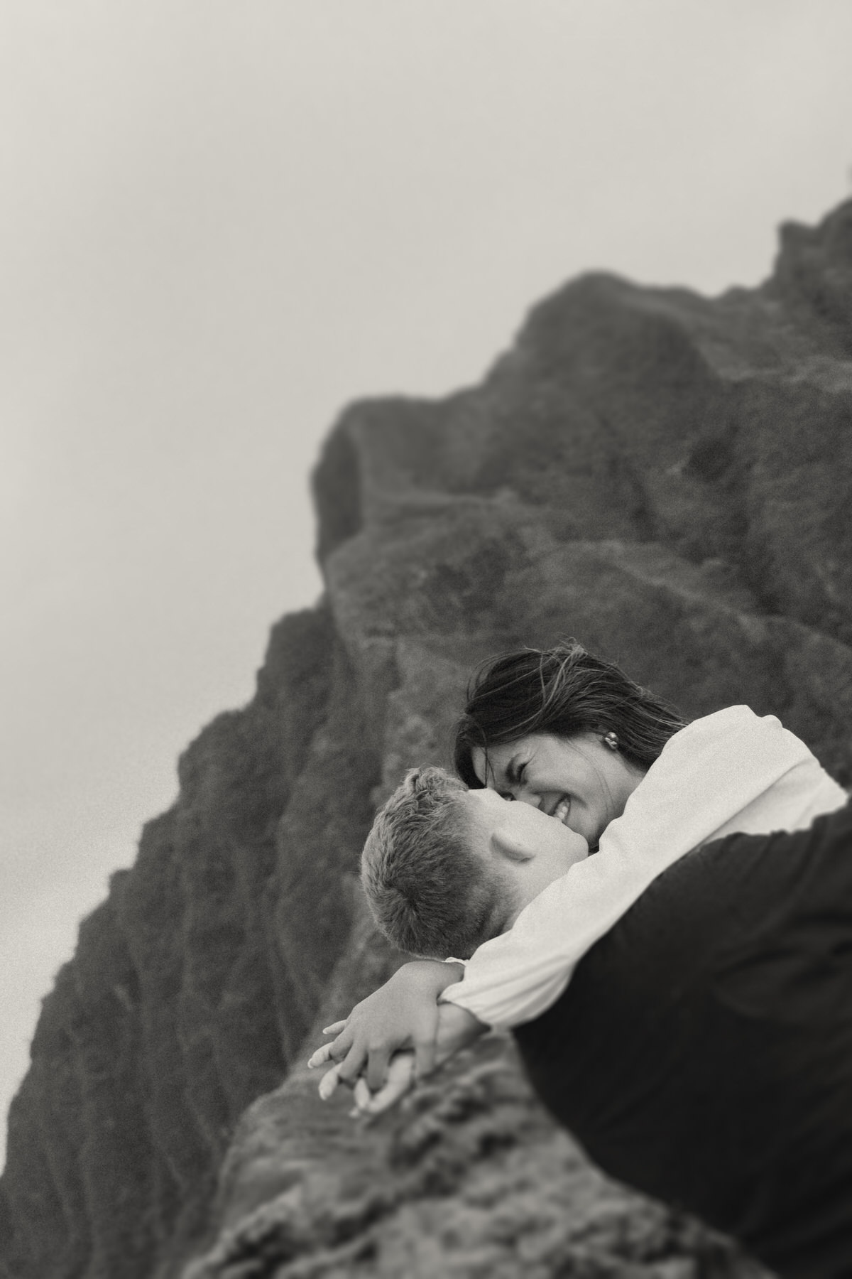 A couple shares a tender moment with one embracing the other, their faces full of joy. The backdrop features dramatic rocky peaks, ideal for adventurous couple photography in Kaneohe's natural environment.