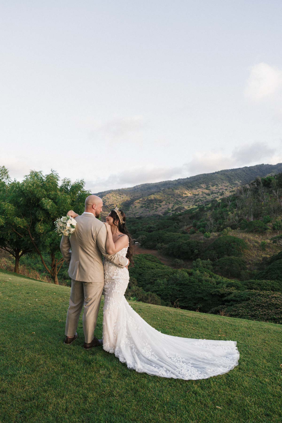 A bride and groom share a quiet moment together in a beautiful open field, with mountains in the distance, perfect for capturing the natural beauty of Kaneohe in wedding photography.