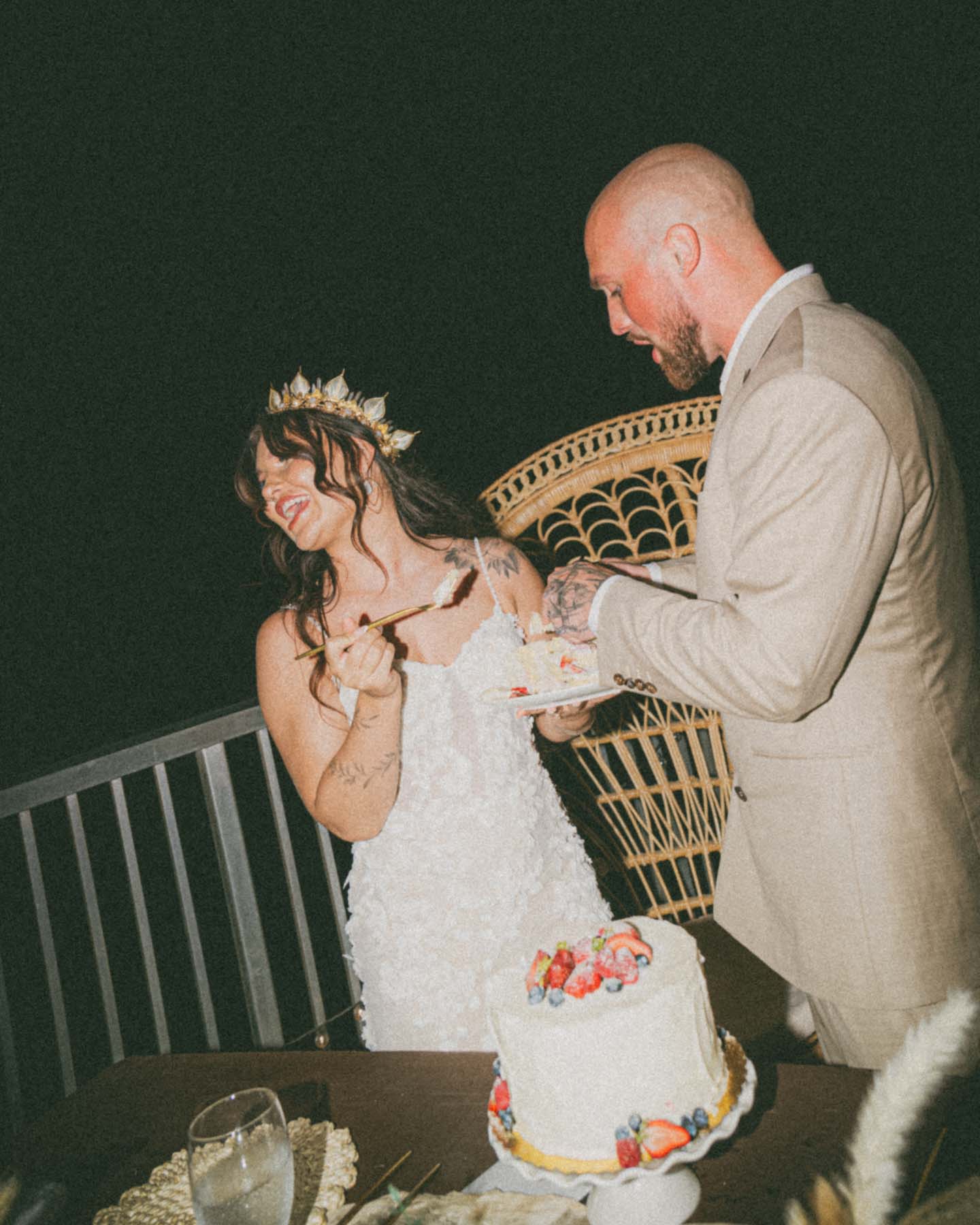 Couple sharing cake at a wedding reception outdoors, with a cake adorned with fresh berries, capturing a playful and affectionate moment between the bride and groom.