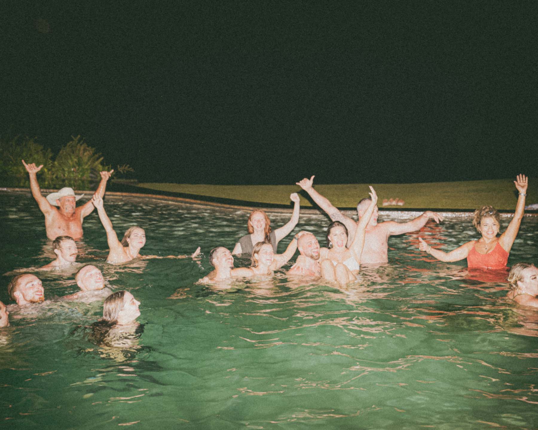 Group of friends celebrating in a pool at night, with their arms raised in joy and laughter, creating an energetic and lively scene at a wedding afterparty.