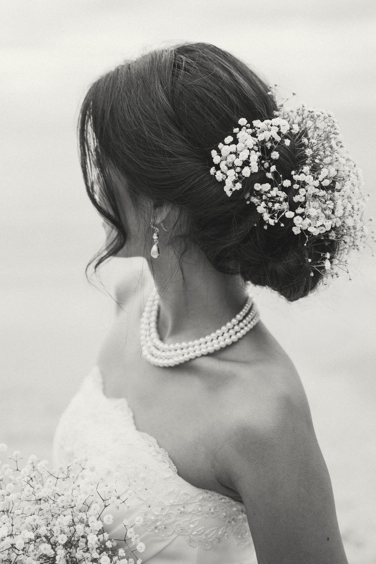 A close-up of a bride's hair and accessories, showing delicate pearls around her neck, earrings, and baby's breath tucked in her hair.