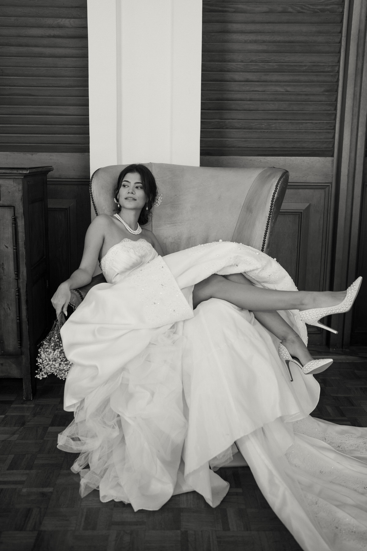 A bride lounges in a vintage armchair, wearing a strapless wedding dress, pearls, and sparkling heels, with a bouquet resting on her lap.