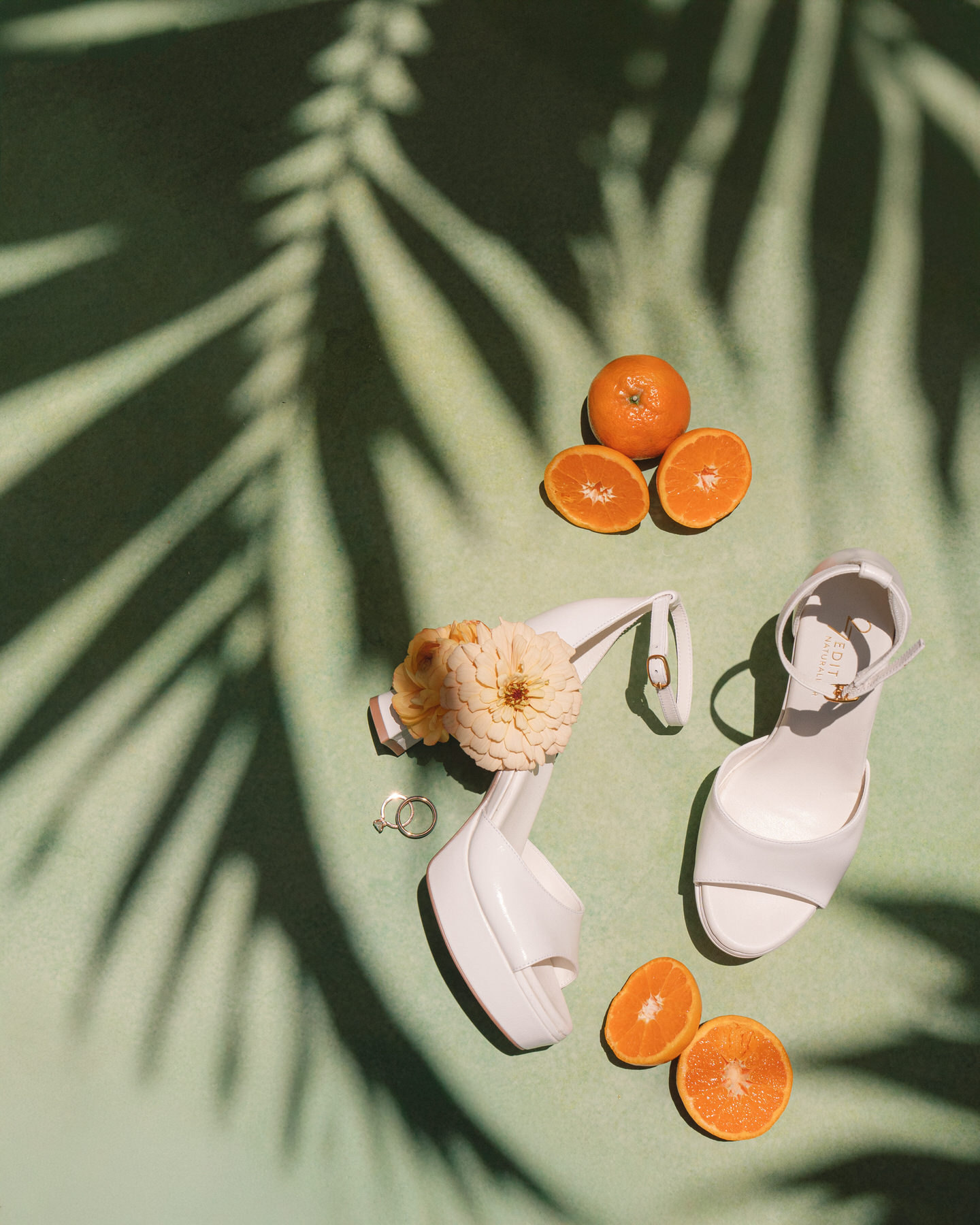 A flat lay of bridal essentials—white block heels, wedding rings, fresh mandarins, and a cream zinnia—styled with tropical shadows for La Pietra Wedding: A Guide for Modern Couples.