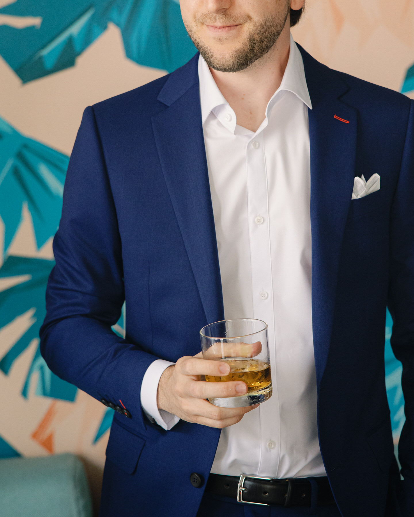A groom in a deep blue suit holds a glass of whiskey, standing against a tropical wallpaper backdrop—sharp style tips from La Pietra Wedding: A Guide for Modern Couples.