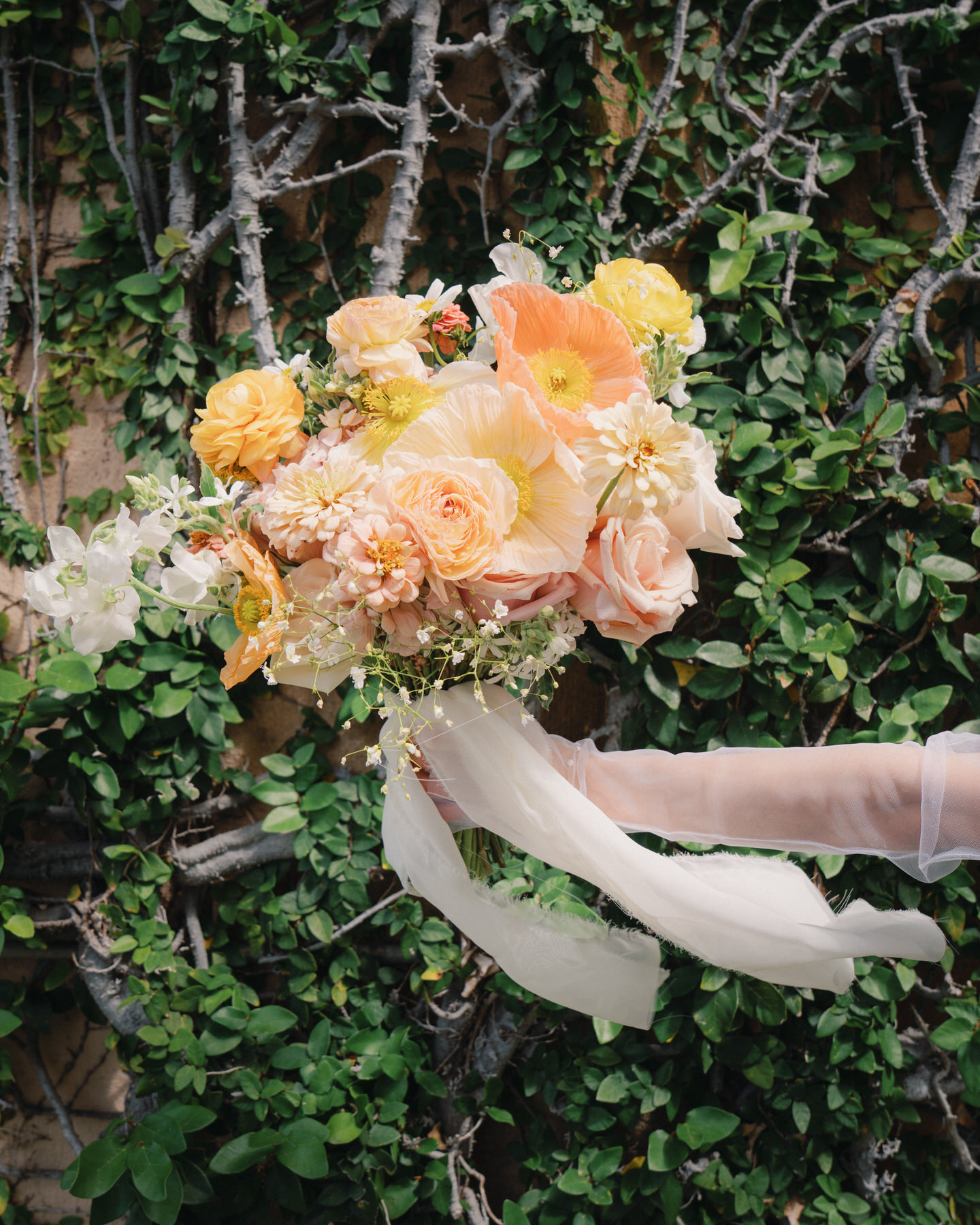 A bride’s arm, dressed in sheer gloves, holds a romantic bouquet of peach, blush, and yellow flowers against a lush vine-covered wall—ideal inspiration from La Pietra Wedding: A Guide for Modern Couples.