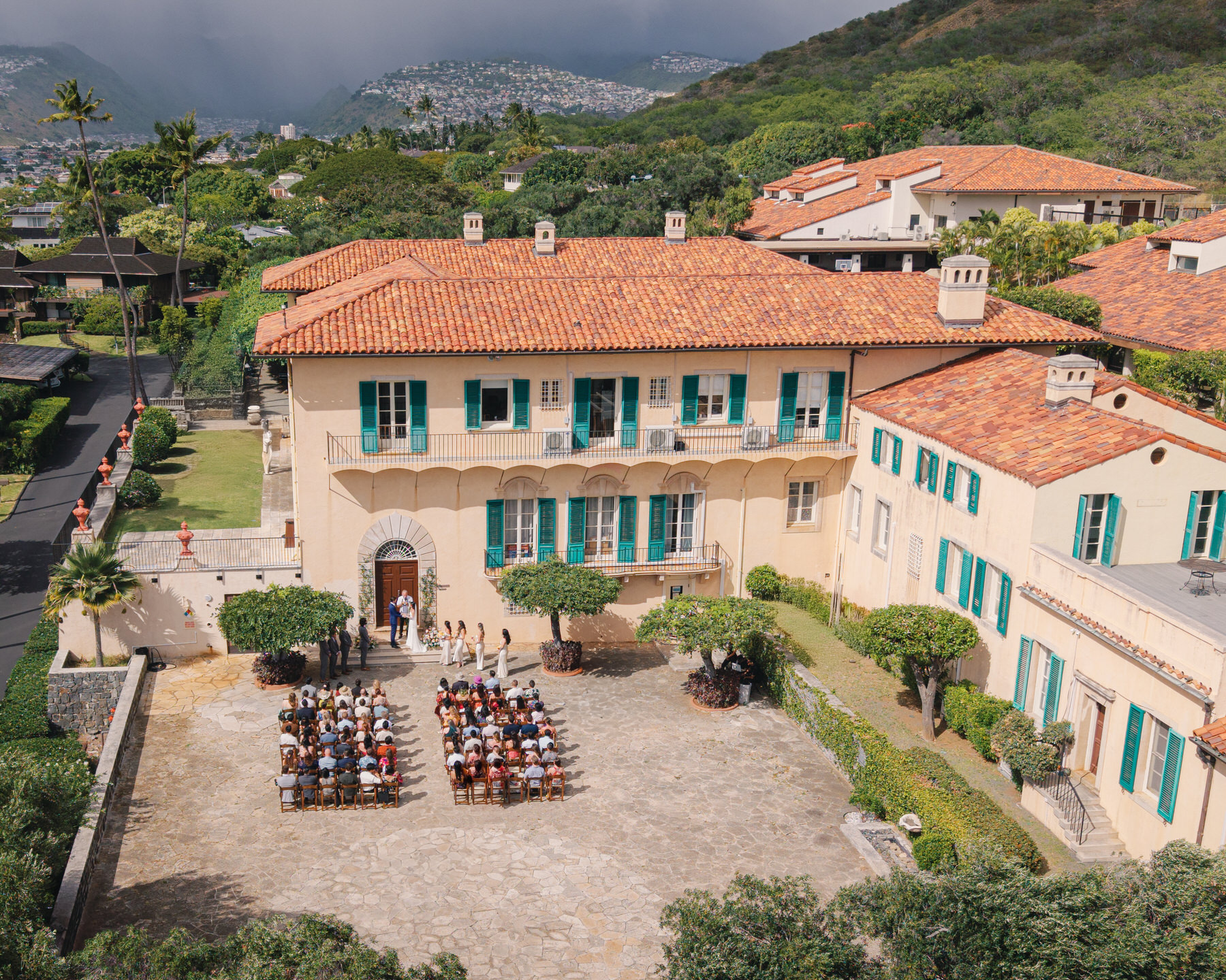 A beautiful outdoor wedding ceremony taking place in front of a grand villa with terracotta-tiled roofs. Rows of guests are seated, with the couple exchanging vows in front of the grand building, surrounded by lush greenery and distant hills.