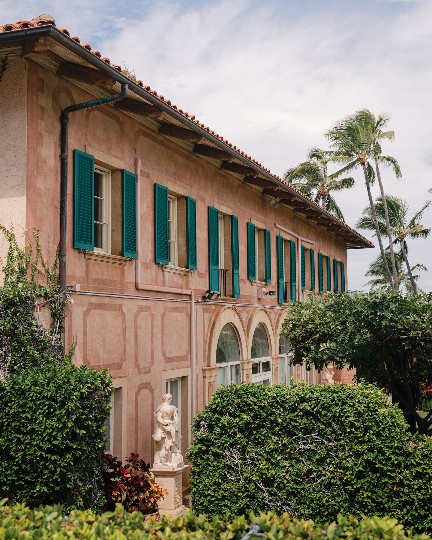 The exterior of La Pietra’s pink-hued villa with green shutters, lush landscaping, and a statue tucked among the foliage—captured for La Pietra Wedding: A Guide for Modern Couples.
