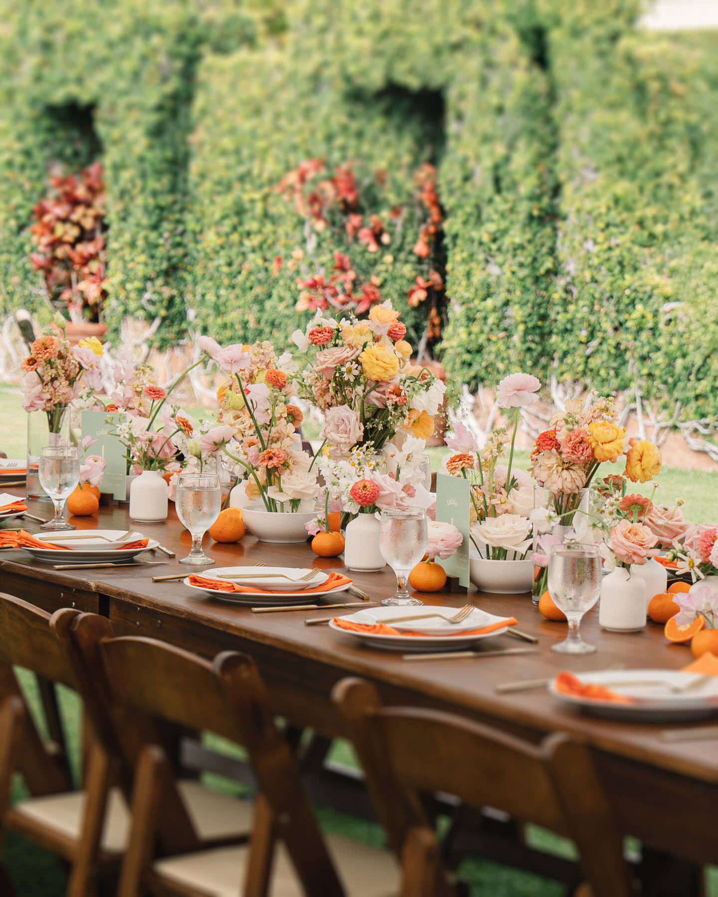A long reception table bursting with colorful floral centerpieces and mandarin oranges, framed by lush green hedges—vibrant styling ideas from La Pietra Wedding: A Guide for Modern Couples.