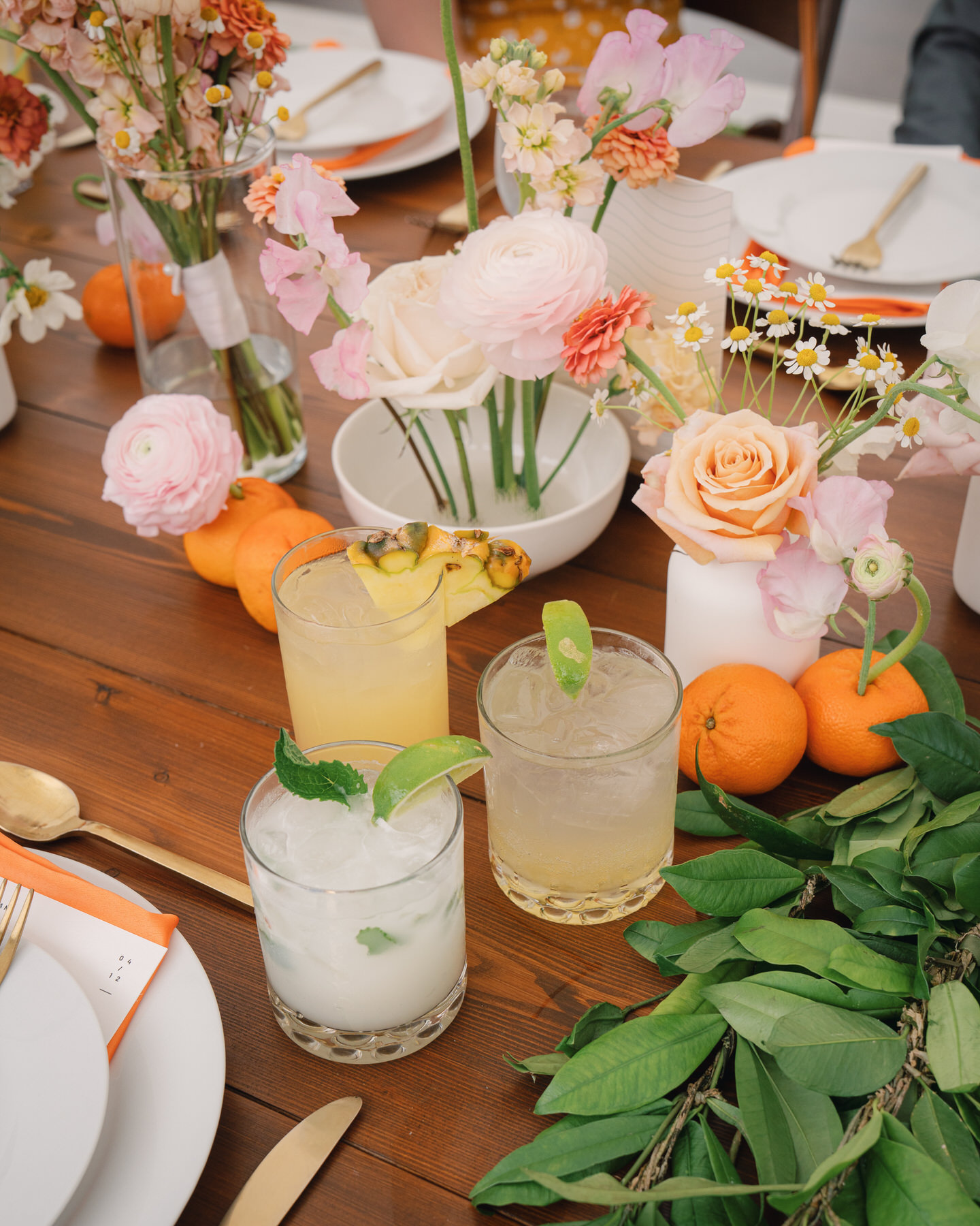 A colorful trio of tropical cocktails surrounded by florals and citrus on a rustic wooden table—refreshing reception details from La Pietra Wedding: A Guide for Modern Couples.