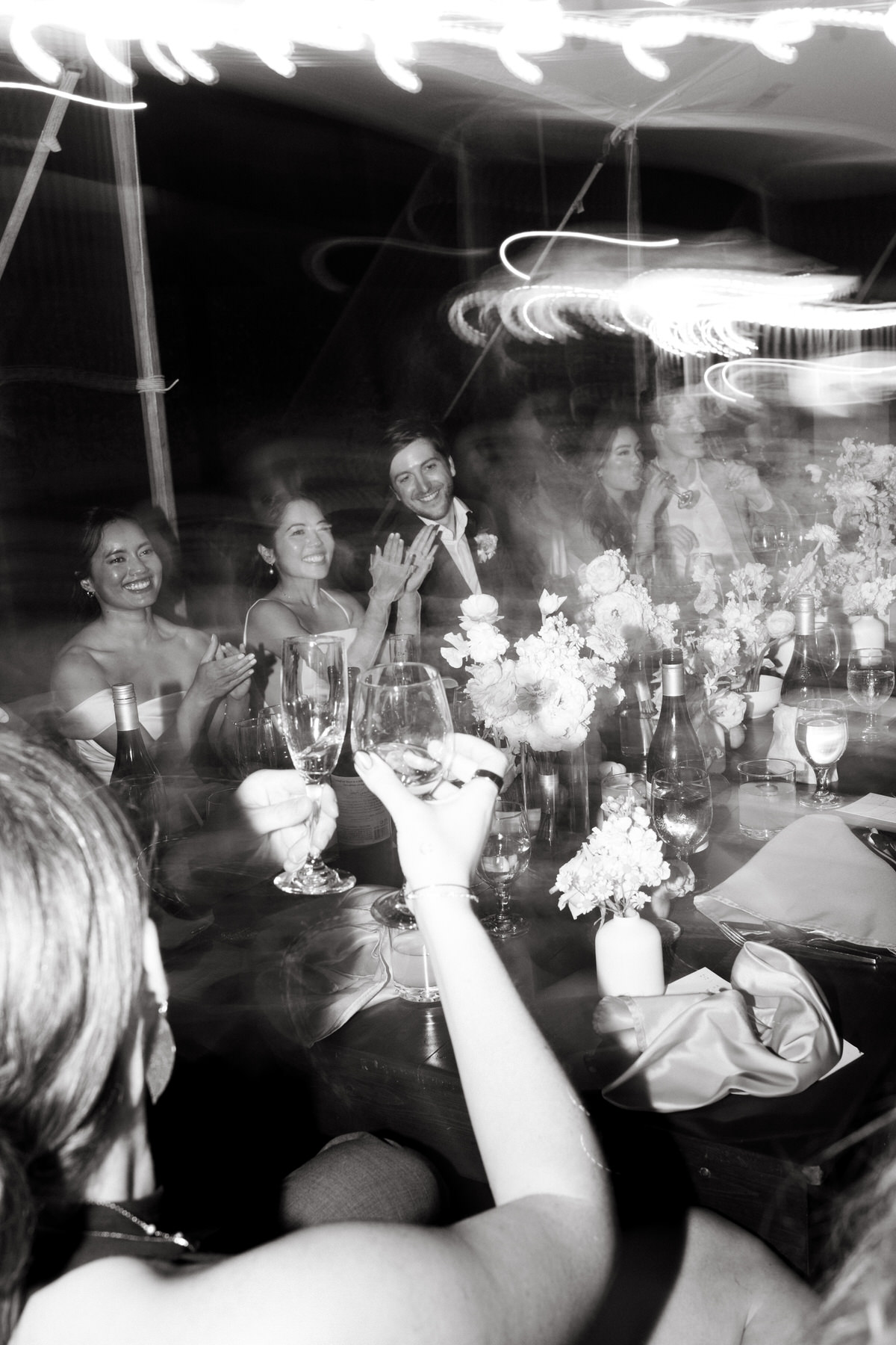 A joyful, motion-blurred black-and-white shot of the newlyweds and guests raising glasses in a nighttime toast—celebration energy in La Pietra Wedding: A Guide for Modern Couples.
