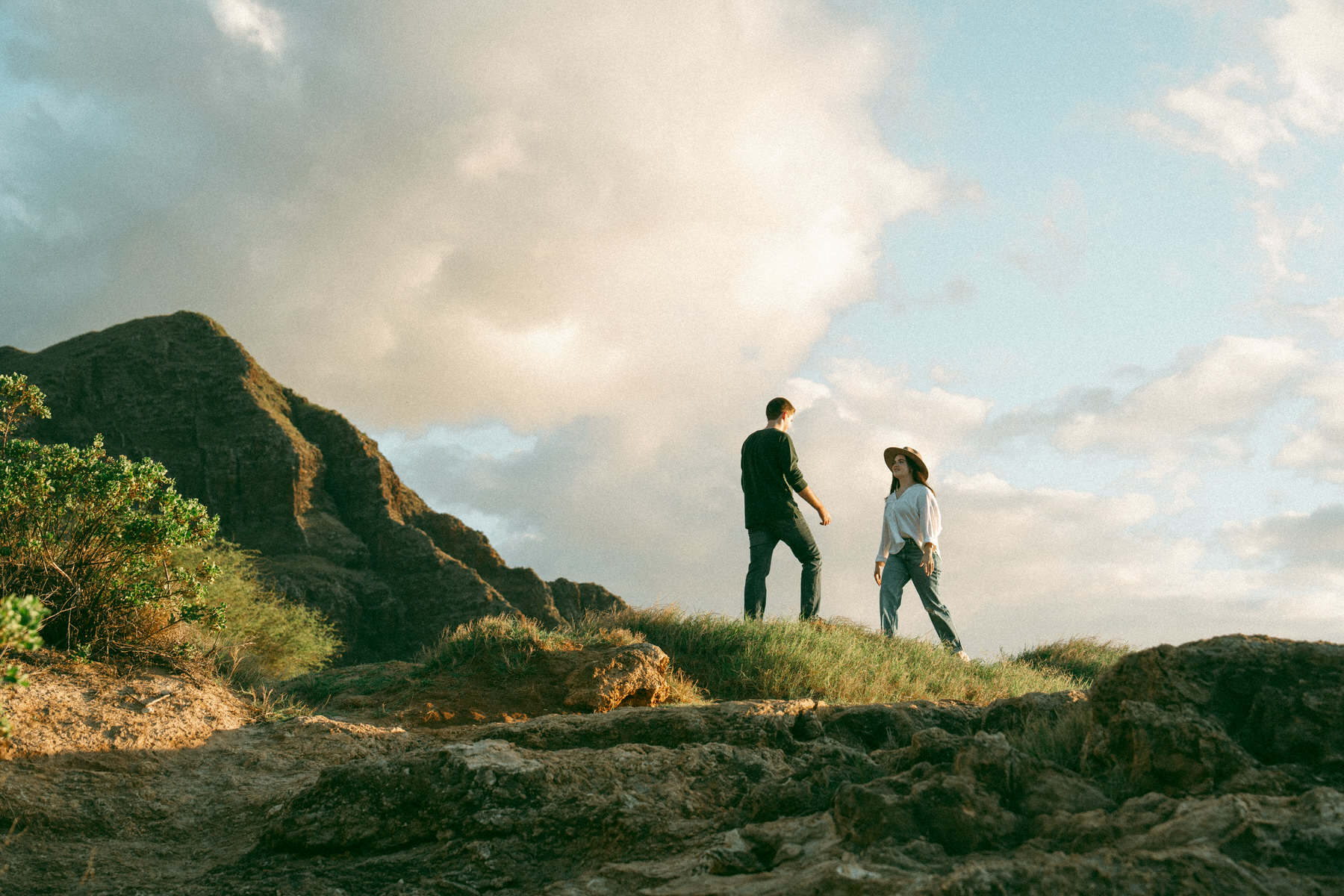 A couple stands on a rocky outcrop overlooking the mountainous terrain, both dressed casually. The woman wears a wide-brimmed hat and smiles as the man extends his hand toward her, with the sun casting a warm glow over the scene.