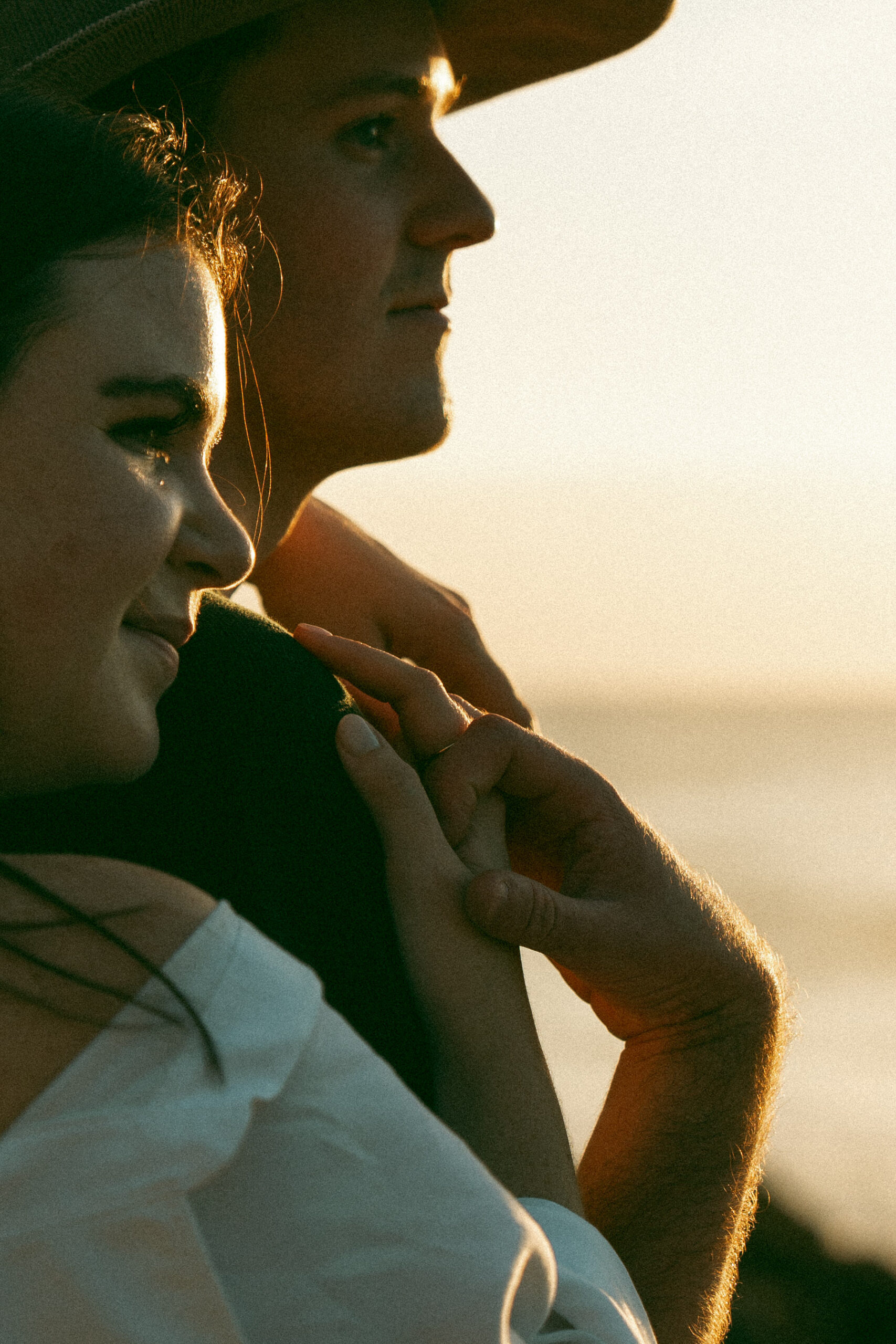 A couple stands close to each other, their faces in profile, as they look toward the horizon during a sunset. The woman, with her hair flowing, smiles softly while the man gazes ahead, his hand resting on her shoulder, creating a peaceful and intimate moment.