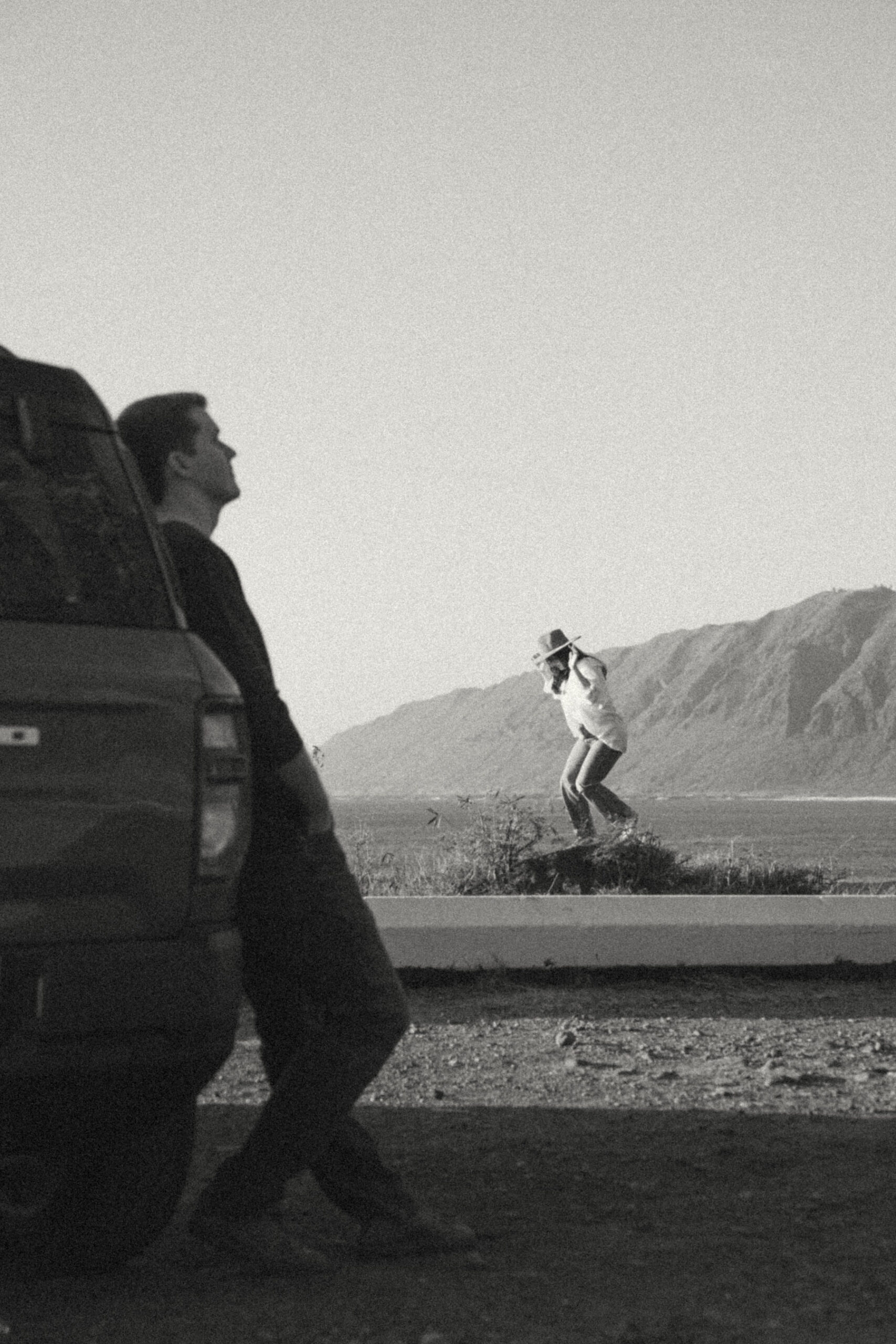 A playful black-and-white image of a woman in a hat jumping in front of a car while the man leans against it, watching her. The man stands still, creating a contrast between the calm and the lively movement of the woman in the background.