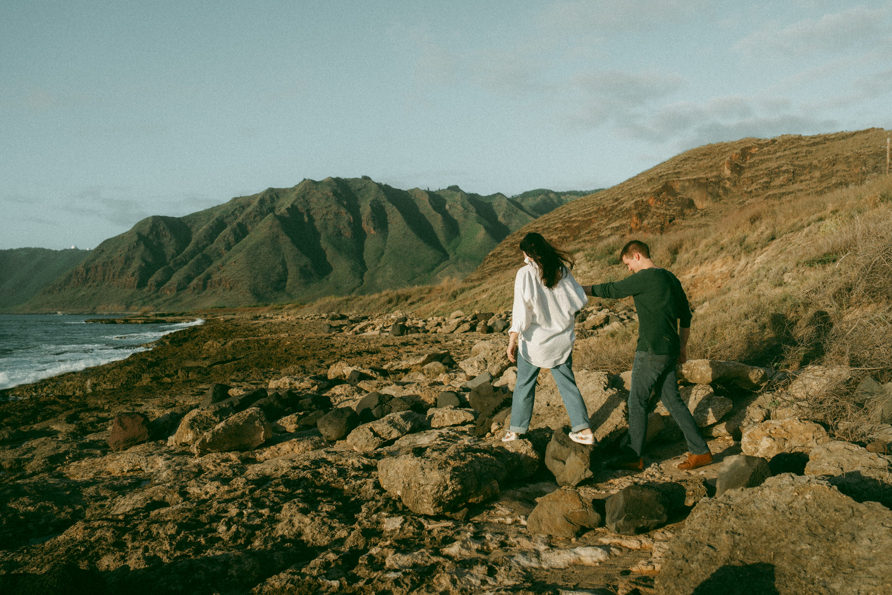 A couple walks hand-in-hand along the rocky shore, the man gently guiding the woman over the rocks. Behind them, green mountains rise above the coastline, and the sky has soft clouds, creating a serene and romantic outdoor scene at Kūhiō Beach