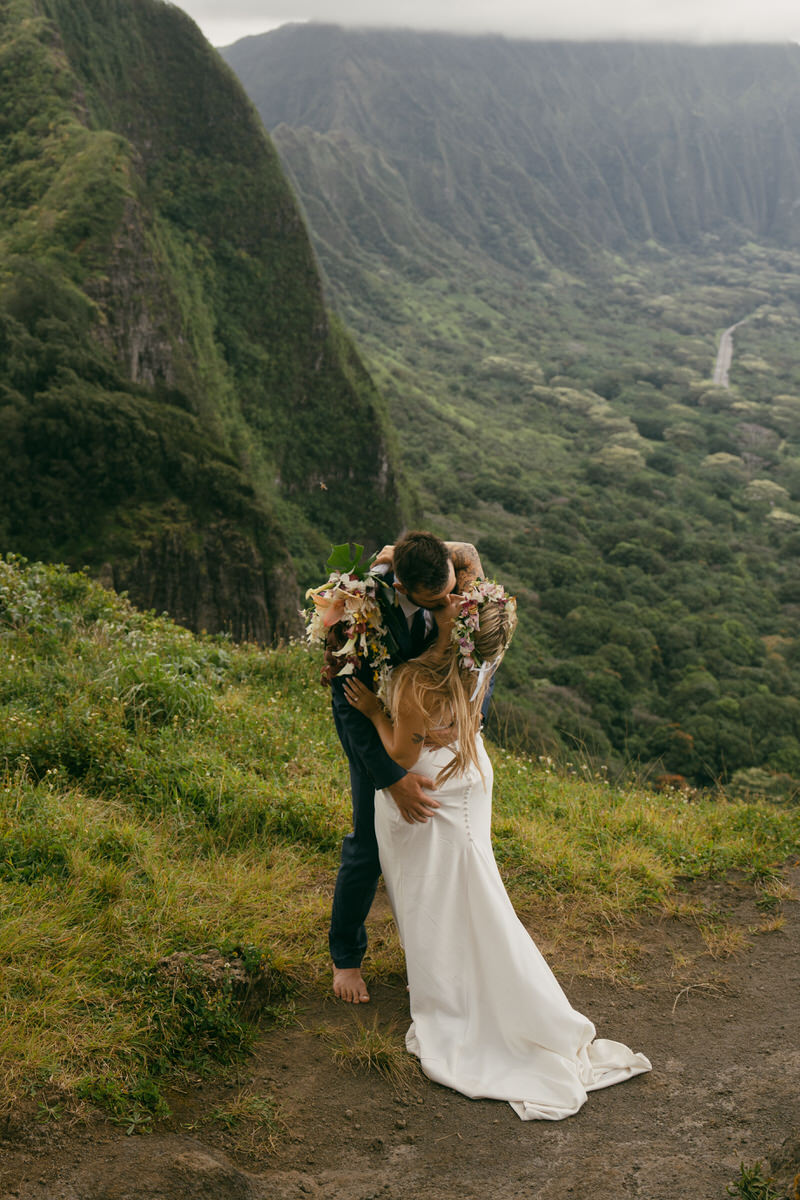 A newlywed couple shares a tender kiss on a lush hillside with dramatic mountain peaks in the background. The bride wears a floral crown, and the groom dons a suit, captured beautifully for a Complete Photography Guide in Kaneohe.