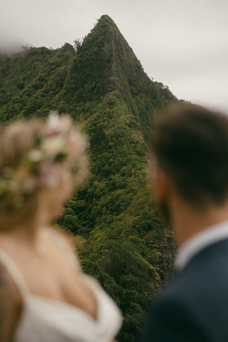 The couple stands in front of a towering, green mountain peak, with the bride gazing off into the distance. Her bouquet and floral crown complement the natural beauty, making this shot perfect for a Complete Photography Guide in Kaneohe.