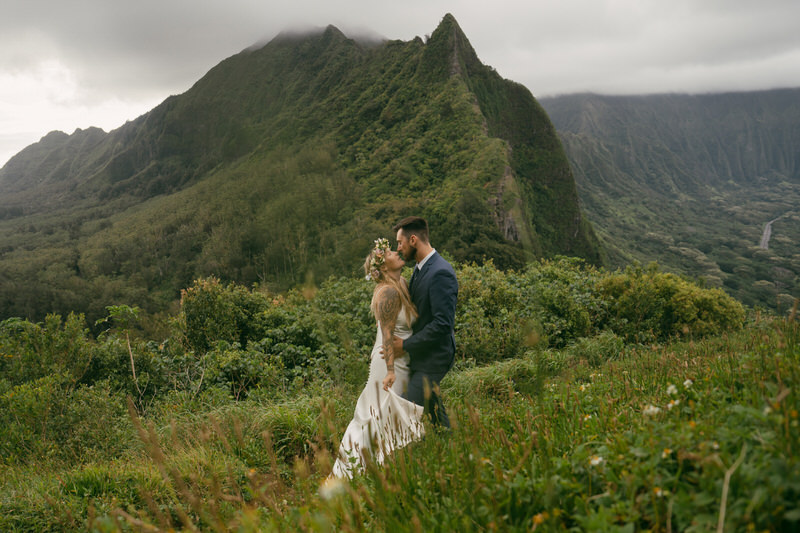 The couple walks hand-in-hand through the lush greenery, with the rugged mountain backdrop adding a sense of adventure. The scene captures a romantic kiss against the dramatic landscape, ideal for a Complete Photography Guide in Kaneohe.