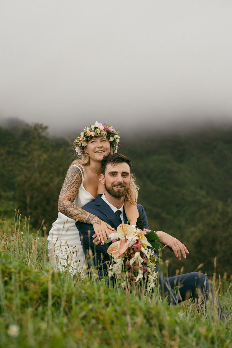 A playful moment with the bride sitting on the groom's lap, both surrounded by vibrant greenery and misty mountains. The groom holds her bouquet as they smile together in this stunning image from a Complete Photography Guide in Kaneohe.
