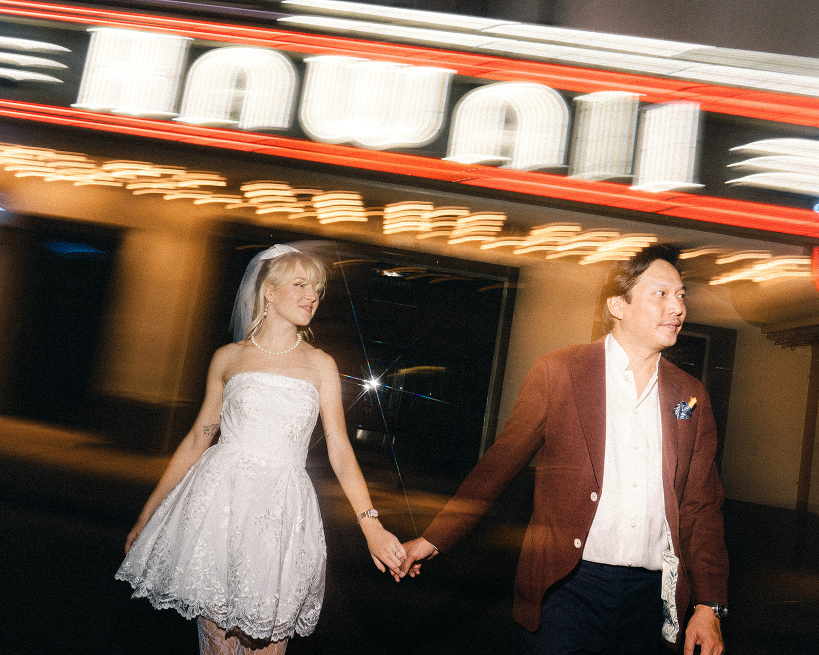 A bride and groom hold hands and walk together under a neon-lit sign in the background, the lights creating motion and adding a playful vibe to the scene.