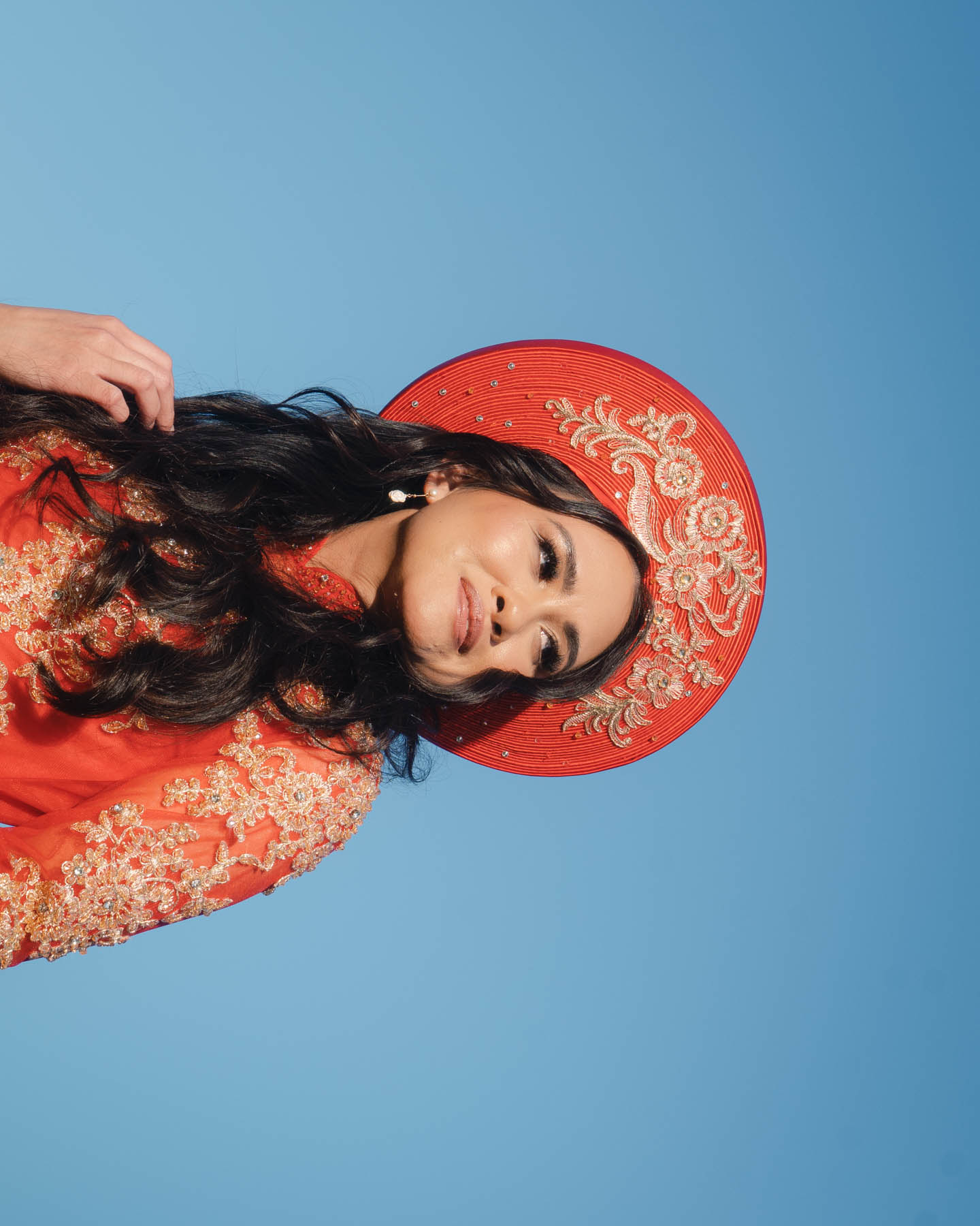 A woman in a bright red, intricately embroidered dress and a matching red hat with golden floral patterns stands against a clear blue sky. Her hair flows gently and she looks to the side.