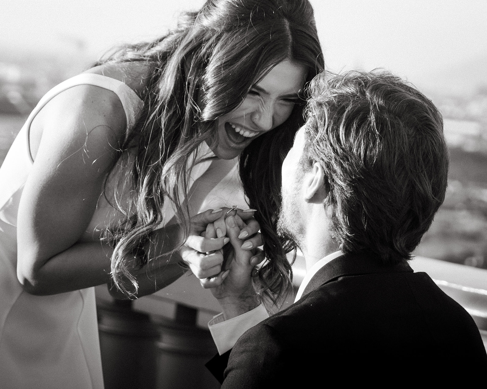 A man in a black suit proposes to a woman in a white dress, both laughing and holding hands with joy in a romantic outdoor setting, captured by an Oahu Proposal Photographer.