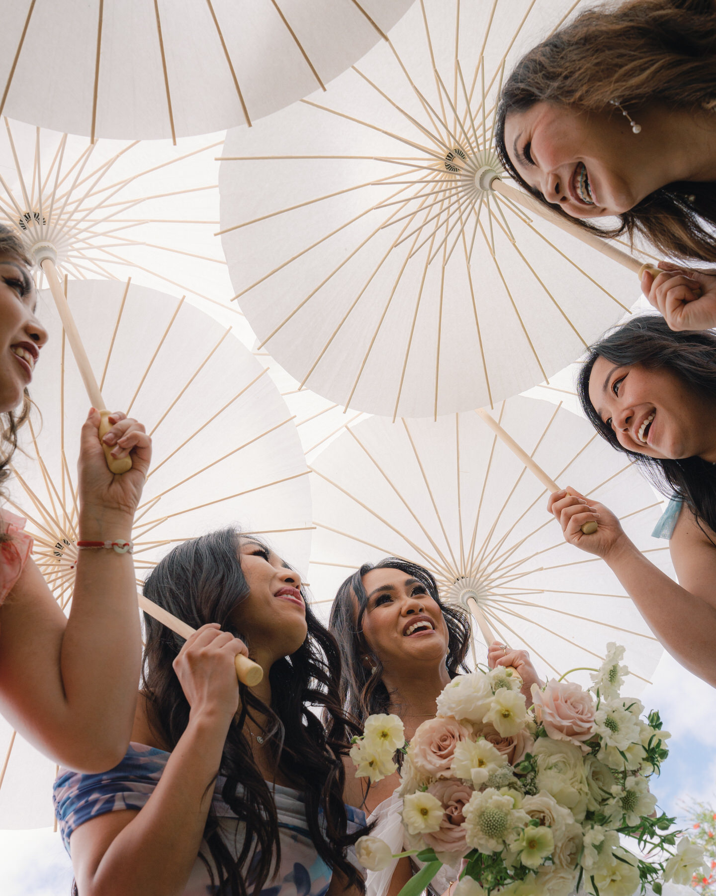 A group of bridesmaids under white parasols, each holding a parasol as they laugh and look up, dressed in various pastel shades and holding floral bouquets.