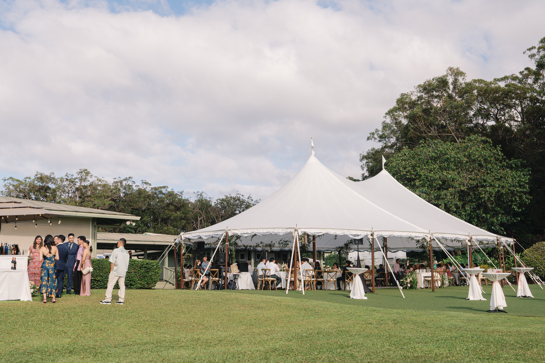 A wedding tent with people gathered outside, sitting at tables and mingling, set in a green grassy field with trees in the background.