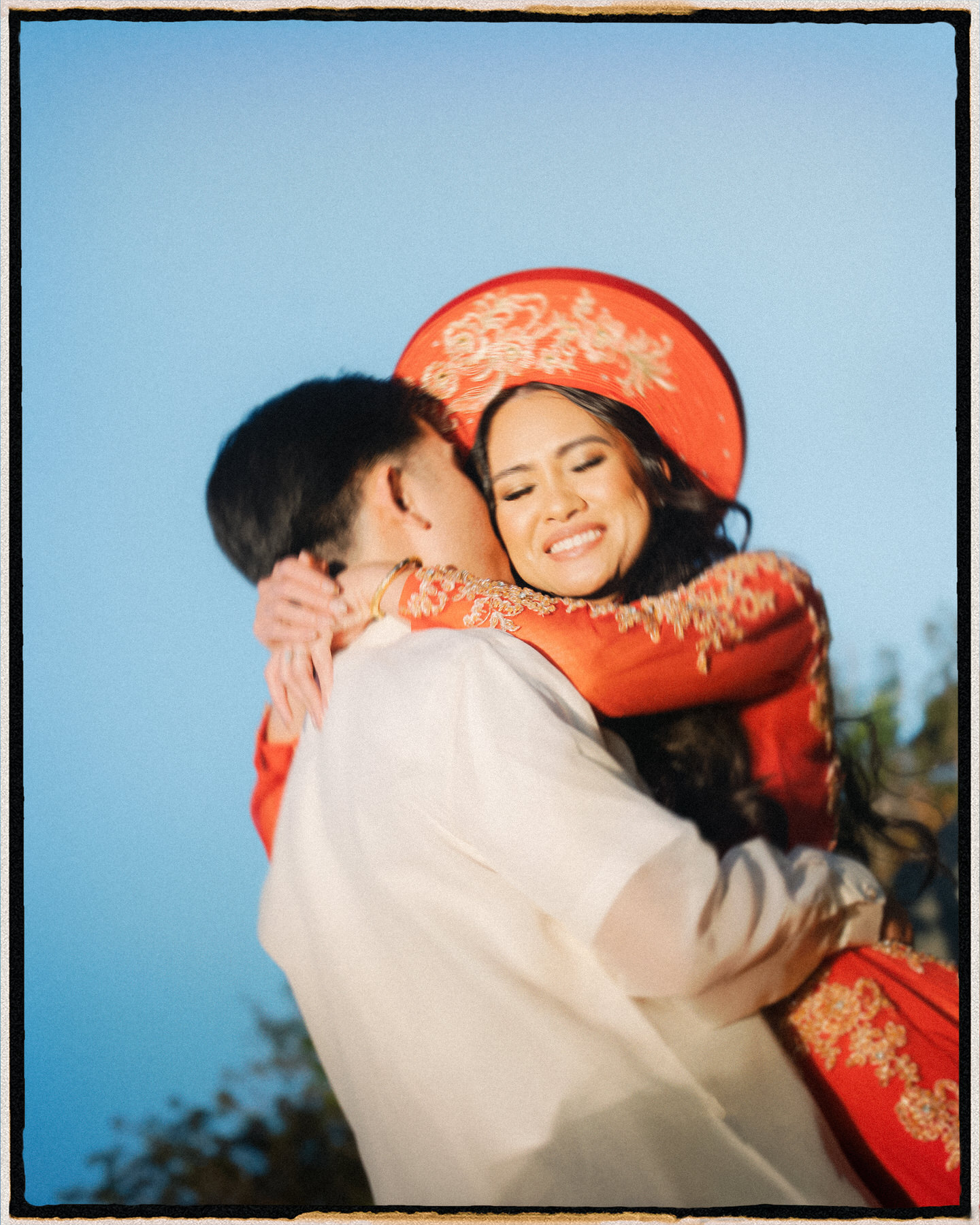 A joyful bride in a red áo dài and khăn đóng hugs her partner at golden hour, blending cultural heritage with love in this moment for a Destination Wedding Photography Guide for Couples Planning a Hawaii Wedding.