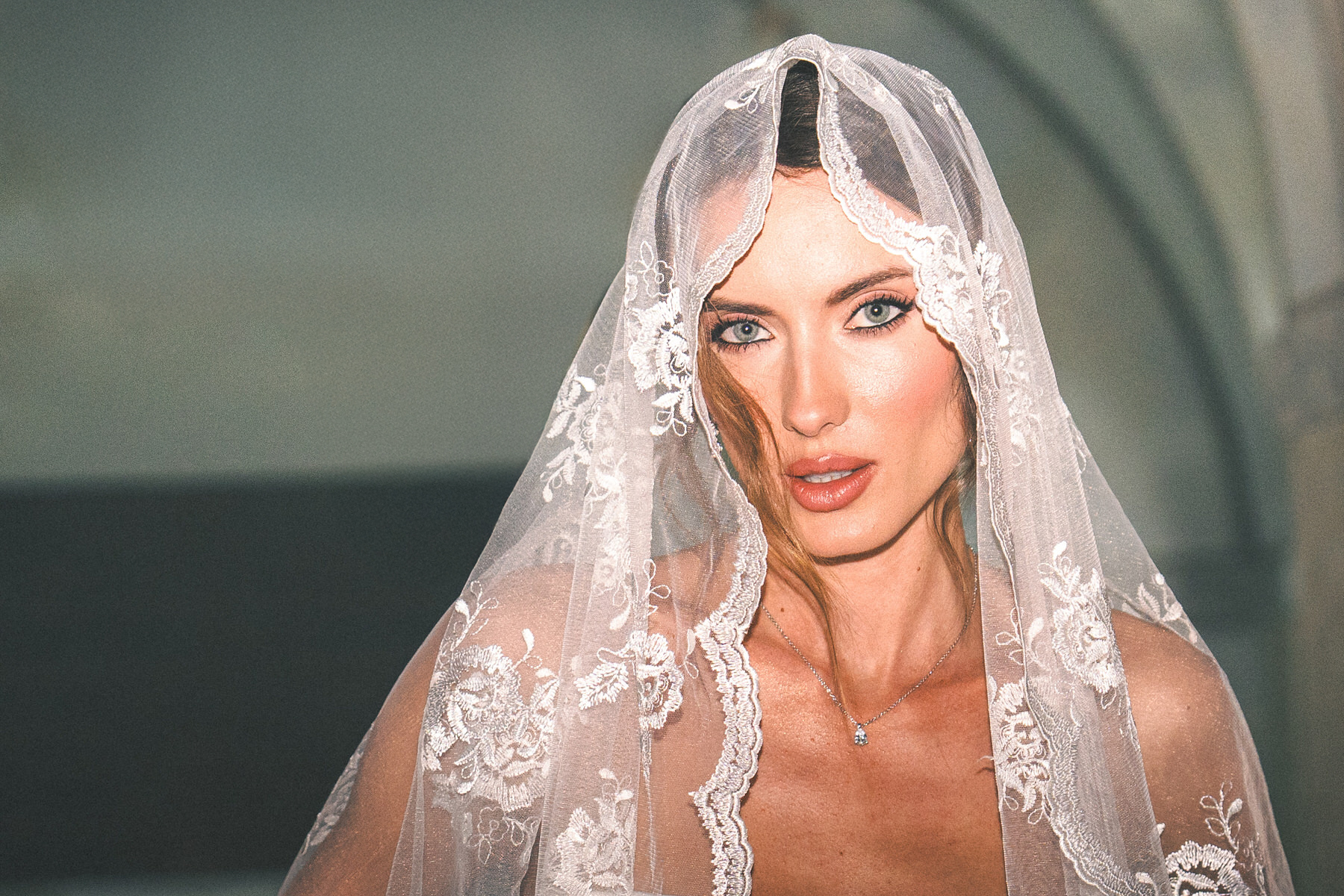 A close-up of a bride with her veil partially covering her face, adorned with intricate lace details. She gazes directly at the camera with a confident and intense expression, showcasing her wedding attire.