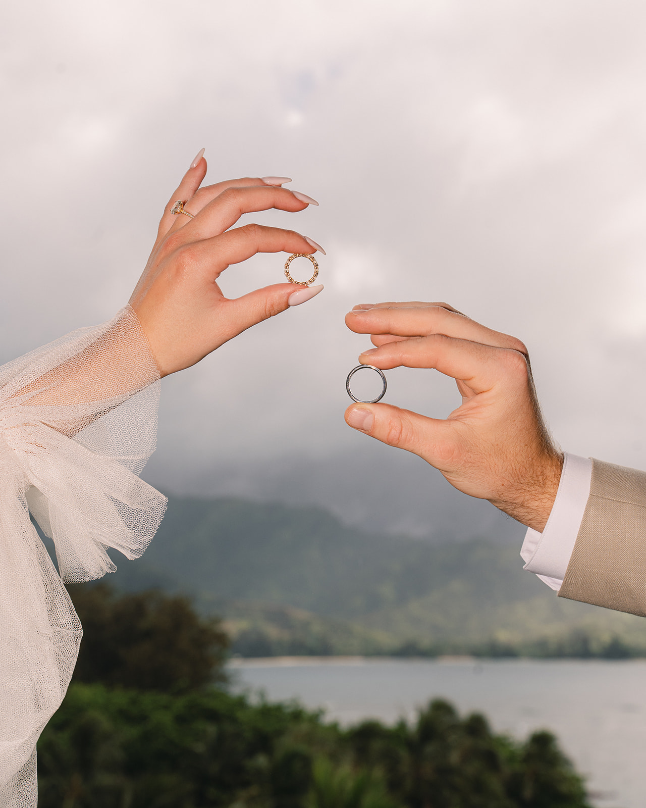 Hands holding wedding rings against mountain and ocean backdrop – Top Questions to Ask Your Wedding Photographer Before Booking