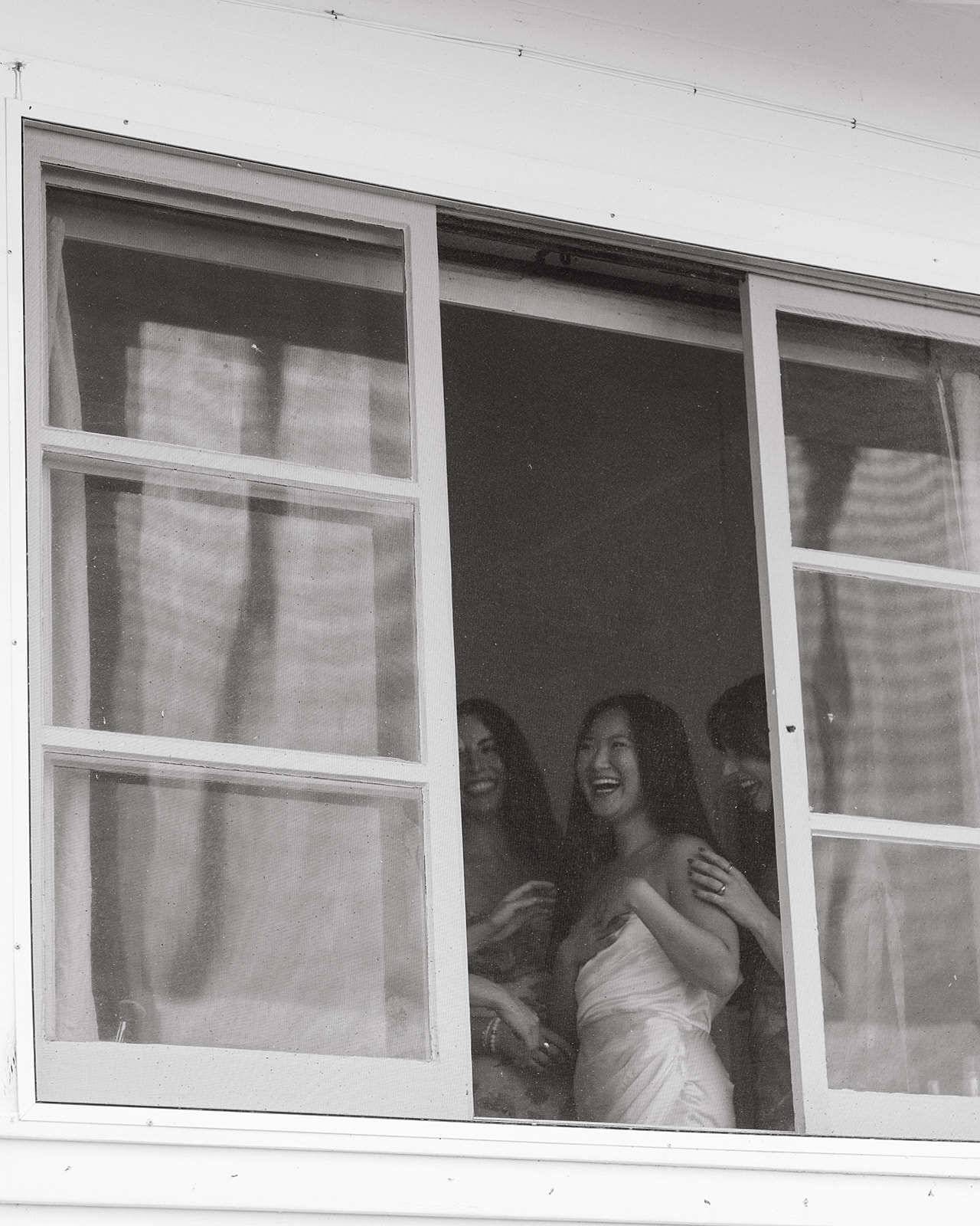 Black-and-white shot of bride and bridesmaids laughing together as they peek out window – How to Choose the Right Photographer for Your Pre-Wedding Shoot