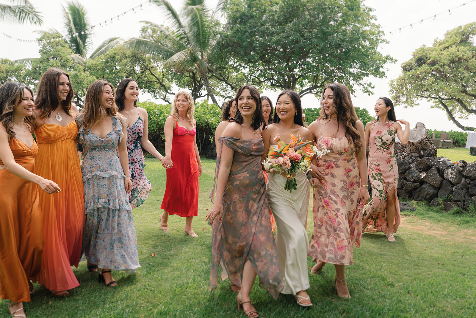 Bride leading her bridesmaids across grassy lawn wearing floral and silk dresses holding bouquet – How to Choose the Right Photographer for Your Pre-Wedding Shoot