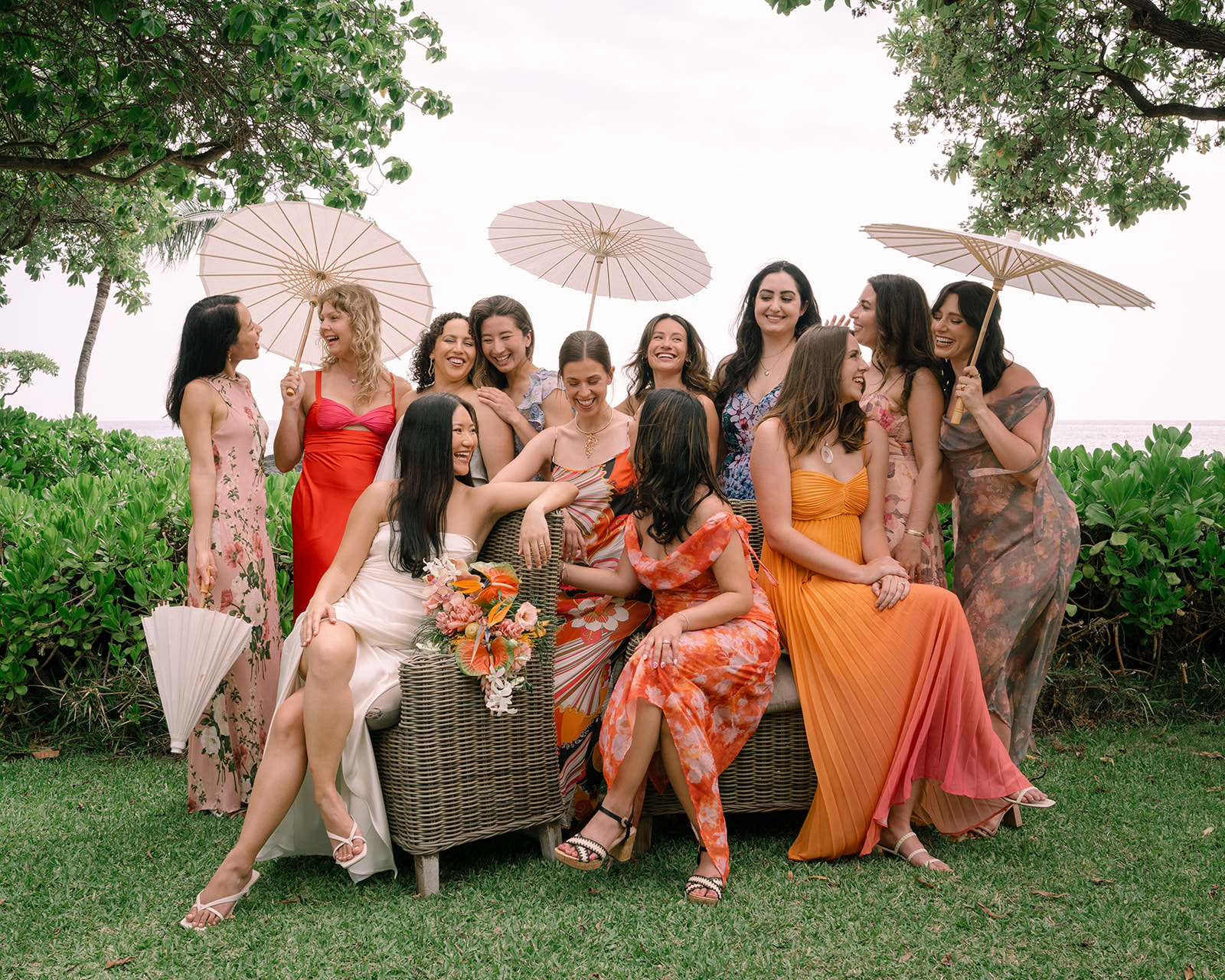 A group of women in colorful dresses and floral crowns gathered outdoors on a wicker couch and chairs, showcasing a joyful moment at hidden gems in Oahu for romantic couple photography