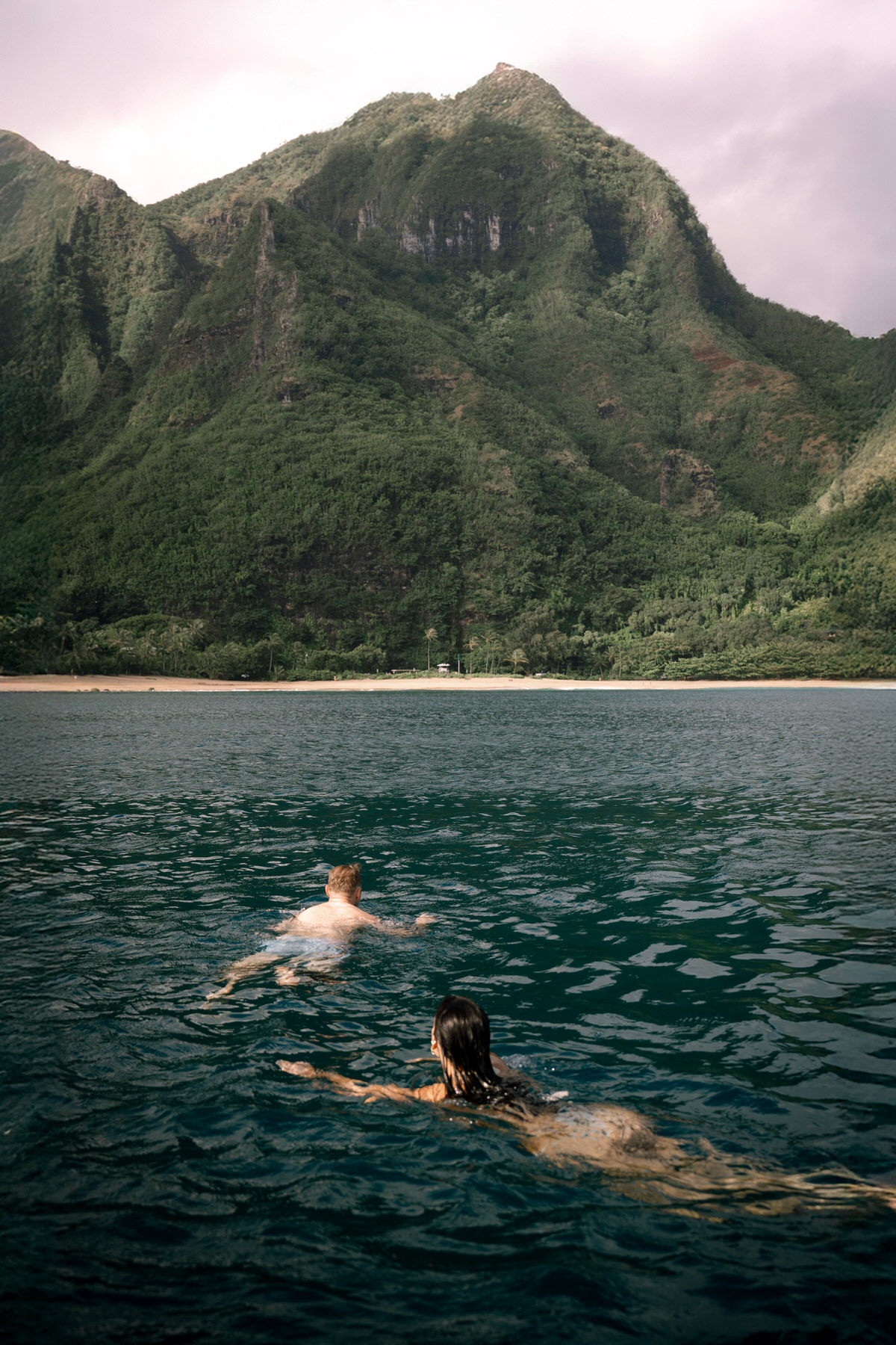 A couple swims in the tranquil waters of Kauai, with lush, dramatic mountains framing the stunning landscape behind them. This image embodies the freedom and intimacy of a Kauai elopement, where couples can connect with nature while enjoying the island's pristine beaches.