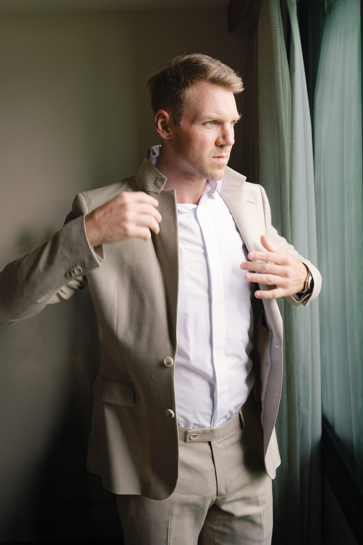 The groom is pictured adjusting his beige suit in front of a window, ready for his Kauai elopement. This moment of final preparation is a key part of planning an intimate and personal elopement, showcasing the relaxed and scenic vibe that Kauai offers couples for their dream elopement.