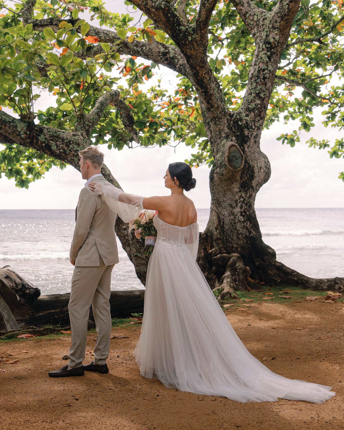 A bride and groom in a romantic first look moment by the beach, as she gently takes taps his shoulder from behind. The stunning Kauai landscape, with the ocean and lush greenery in the background, creates an intimate atmosphere, perfect for planning your dream Kauai elopement.