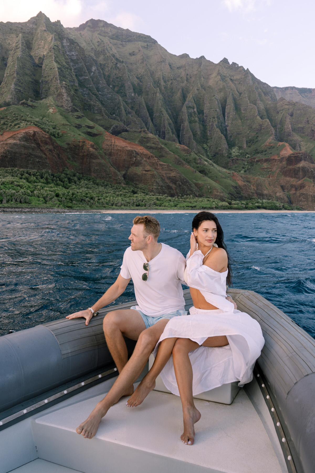 A couple sits on a boat in Kauai, surrounded by majestic mountain views. As part of your dream Kauai elopement, you can embrace the island's natural beauty by incorporating scenic boat rides into your wedding experience, offering breathtaking backdrops for your special day.