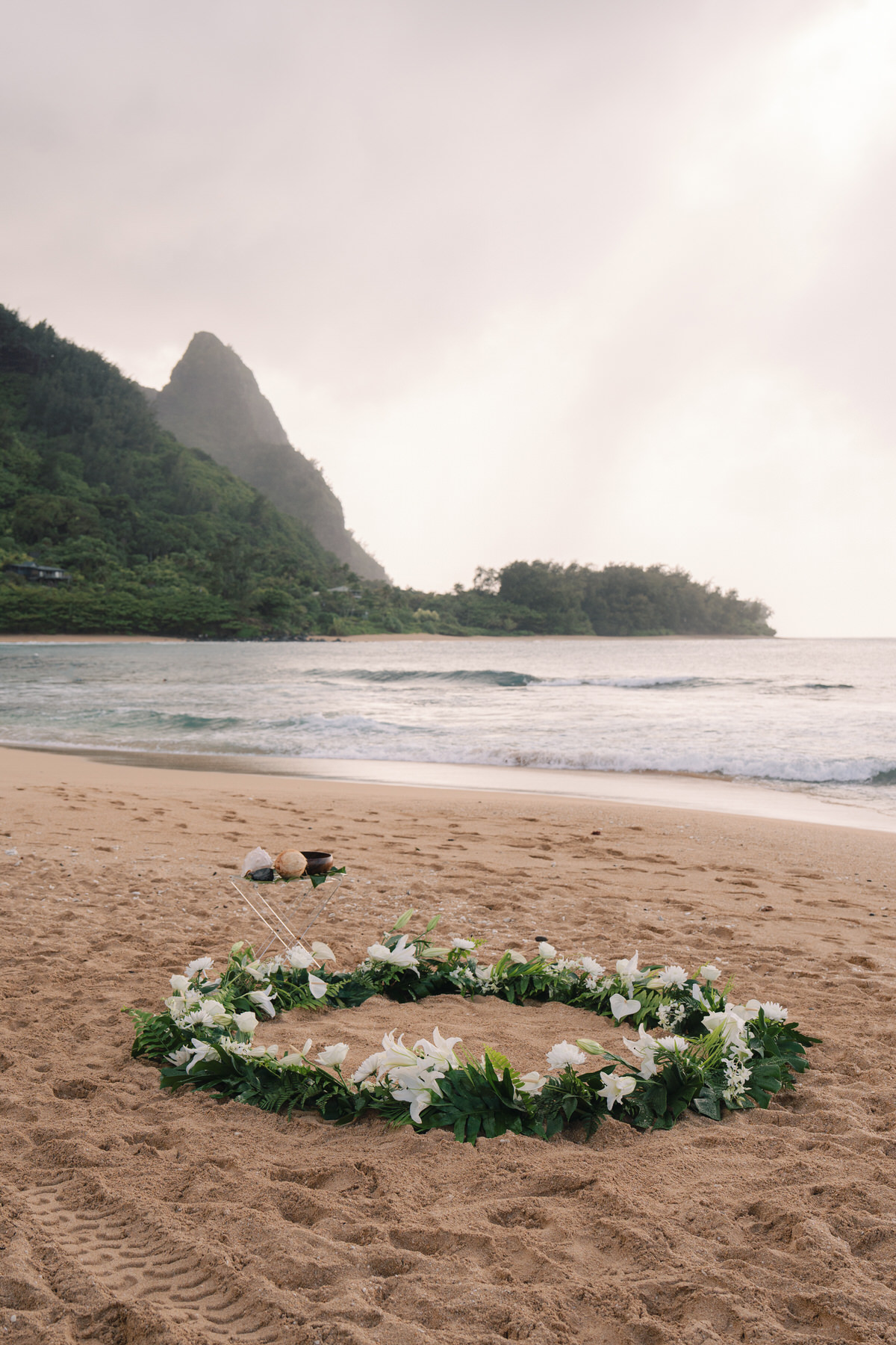 A circular floral arrangement, made with white lilies and greenery, sits on the sand near the water’s edge, creating a romantic and serene setting for a Kauai elopement ceremony. The backdrop of mountains and the ocean emphasizes the importance of choosing the perfect location for your elopement.