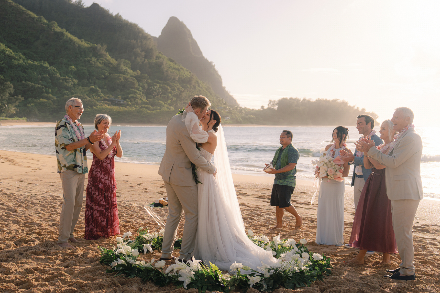 The bride and groom share a passionate kiss during their Kauai elopement ceremony on the beach. Their ceremony is framed by lush greenery and the dramatic coastline, capturing the essence of a Kauai elopement—simple yet breathtaking, with close family or friends in attendance. The soft sunset light adds to the magical and intimate atmosphere of the day.
