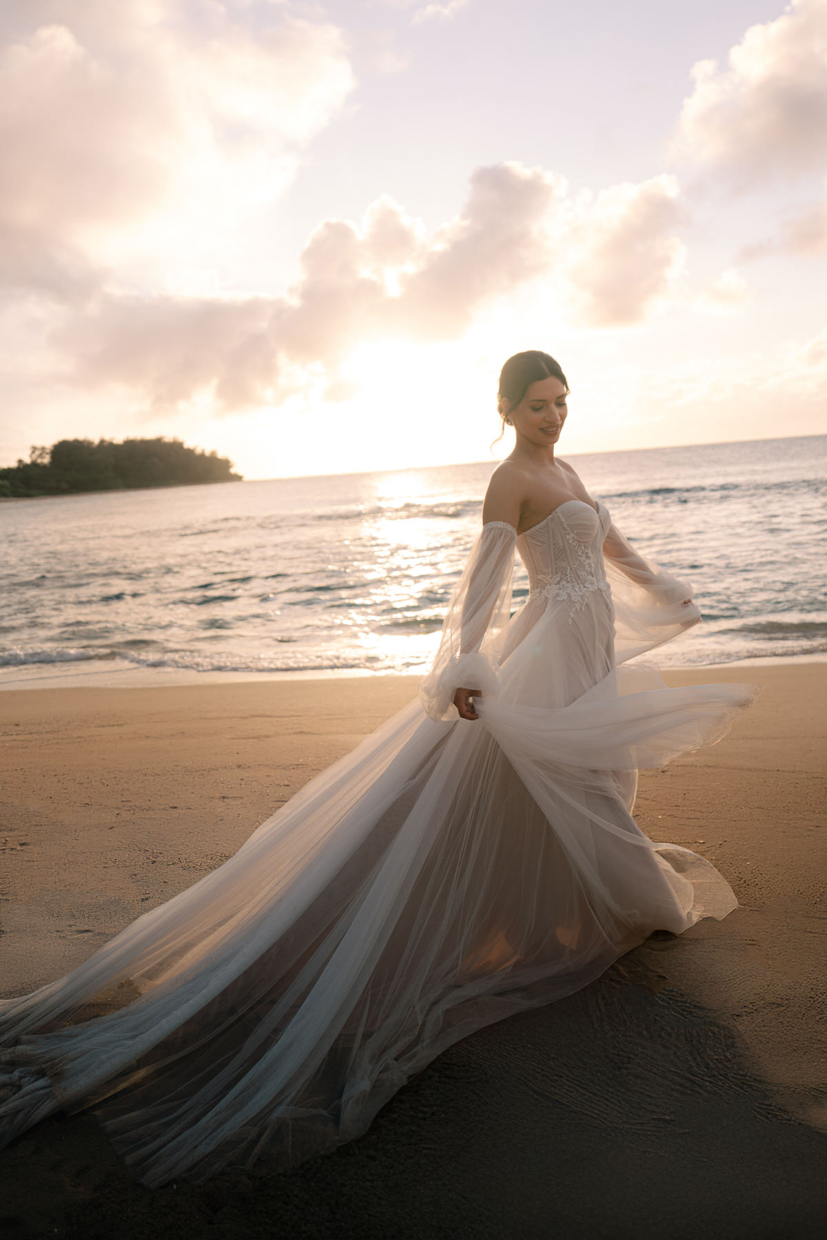 The bride walks along the beach, her wedding gown flowing in the breeze, while the sun sets behind her. The stunning Kauai scenery serves as the perfect backdrop for this dream Kauai elopement moment, capturing the essence of love and nature coming together.