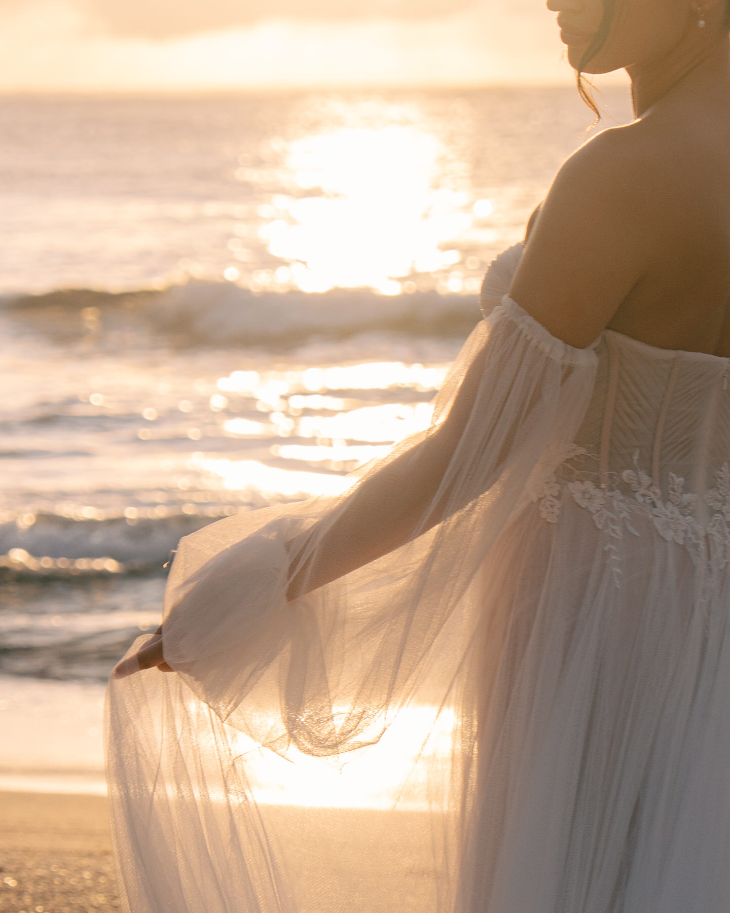 The bride stands on the beach at sunset, her flowing wedding gown catching the light of the sun as she delicately holds her dress. This serene scene captures the essence of an idyllic Kauai elopement, where natural beauty and simplicity combine perfectly for a magical moment.